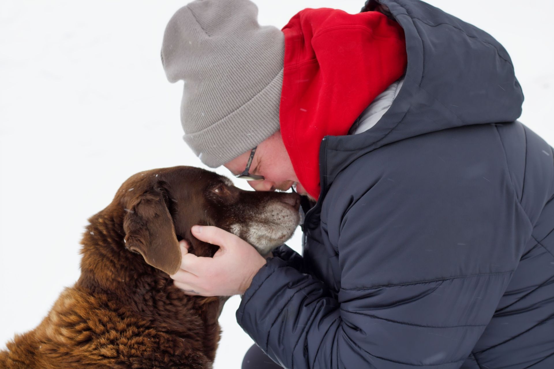 Person in blue coat and hat kisses brown dog's snout in snowy setting.