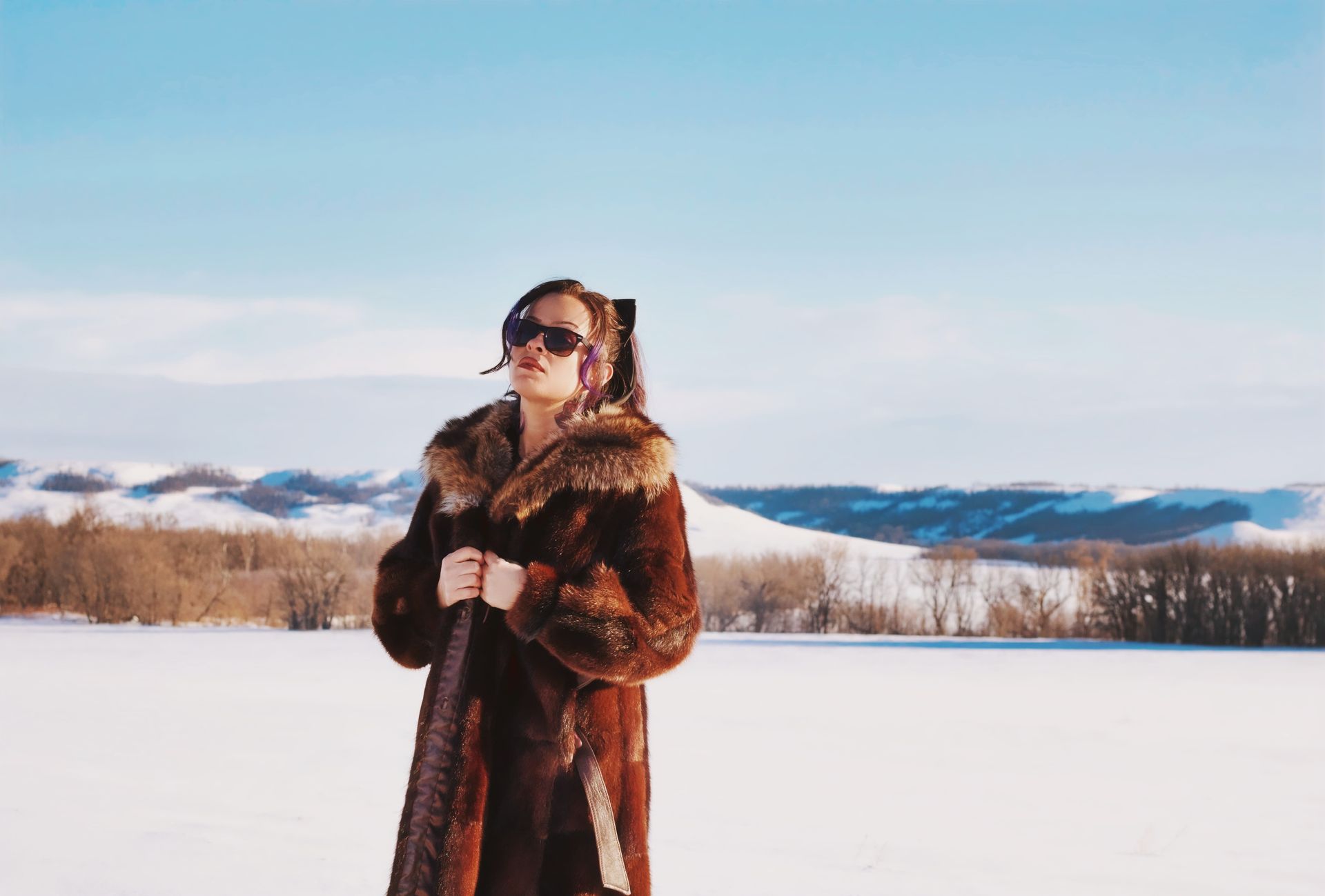 Woman in a brown fur coat and sunglasses standing in a snowy field on a sunny day.