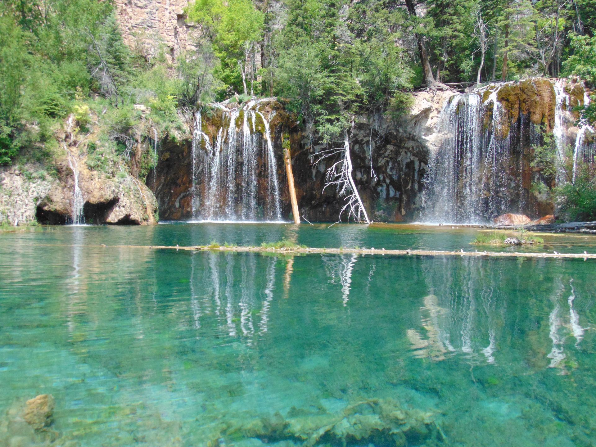 Crystal clear turquoise lake with multiple waterfalls cascading over a rock face, surrounded by trees.