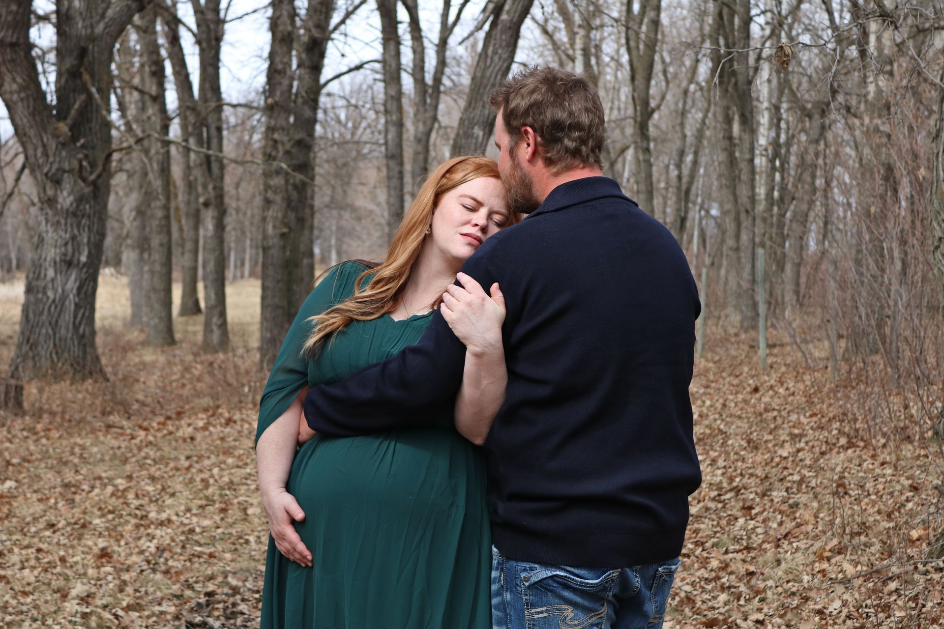 A pregnant person in a long green dress and a partner embrace in a wooded area with fallen leaves on the ground.