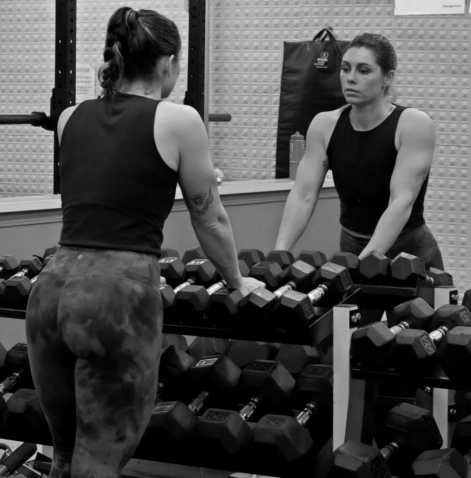 Woman in gym outfit looking at reflection in mirror, dumbbells in foreground.