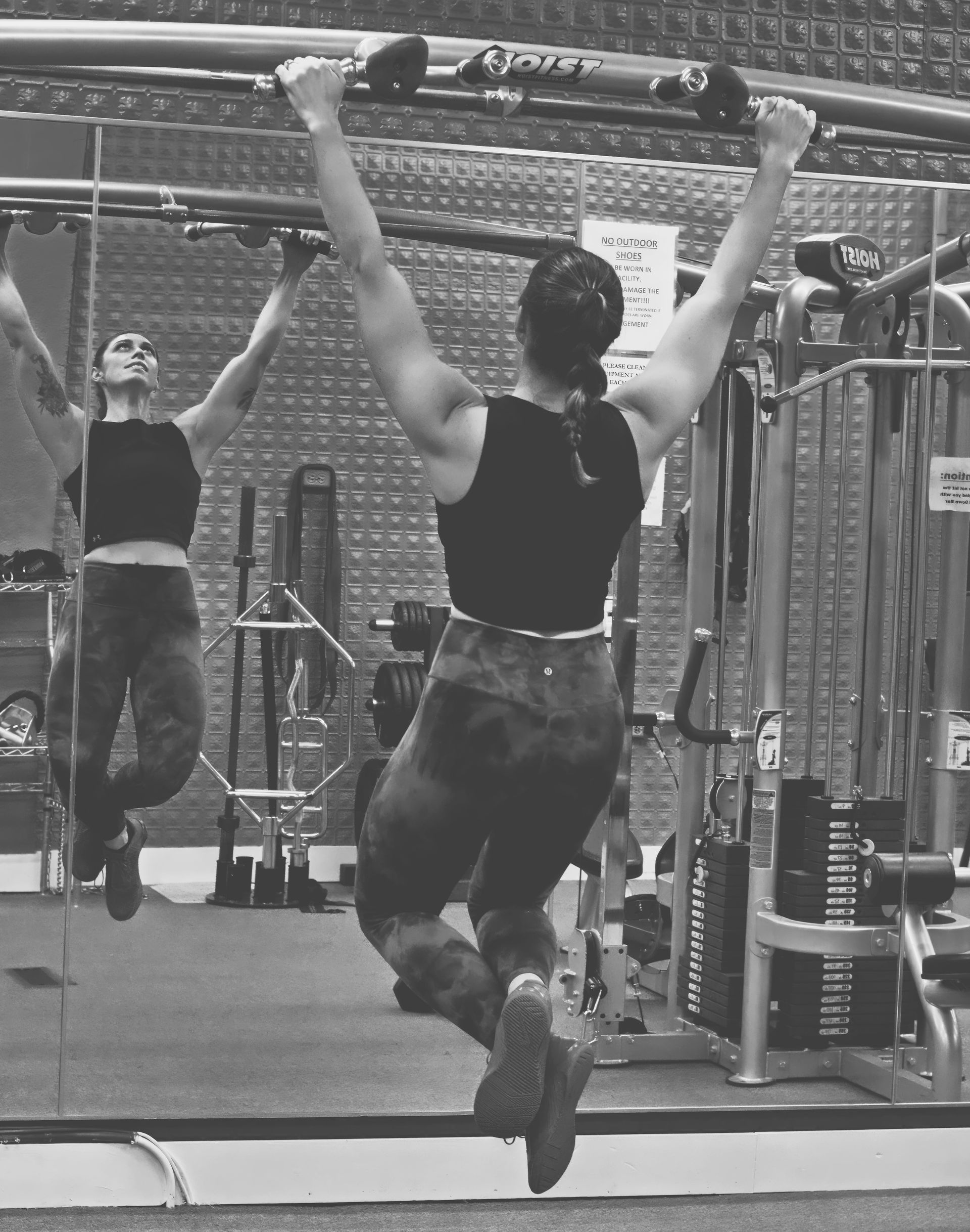 Woman doing a pull-up in a gym, reflected in a mirror. She's wearing workout clothes, with arms fully extended.