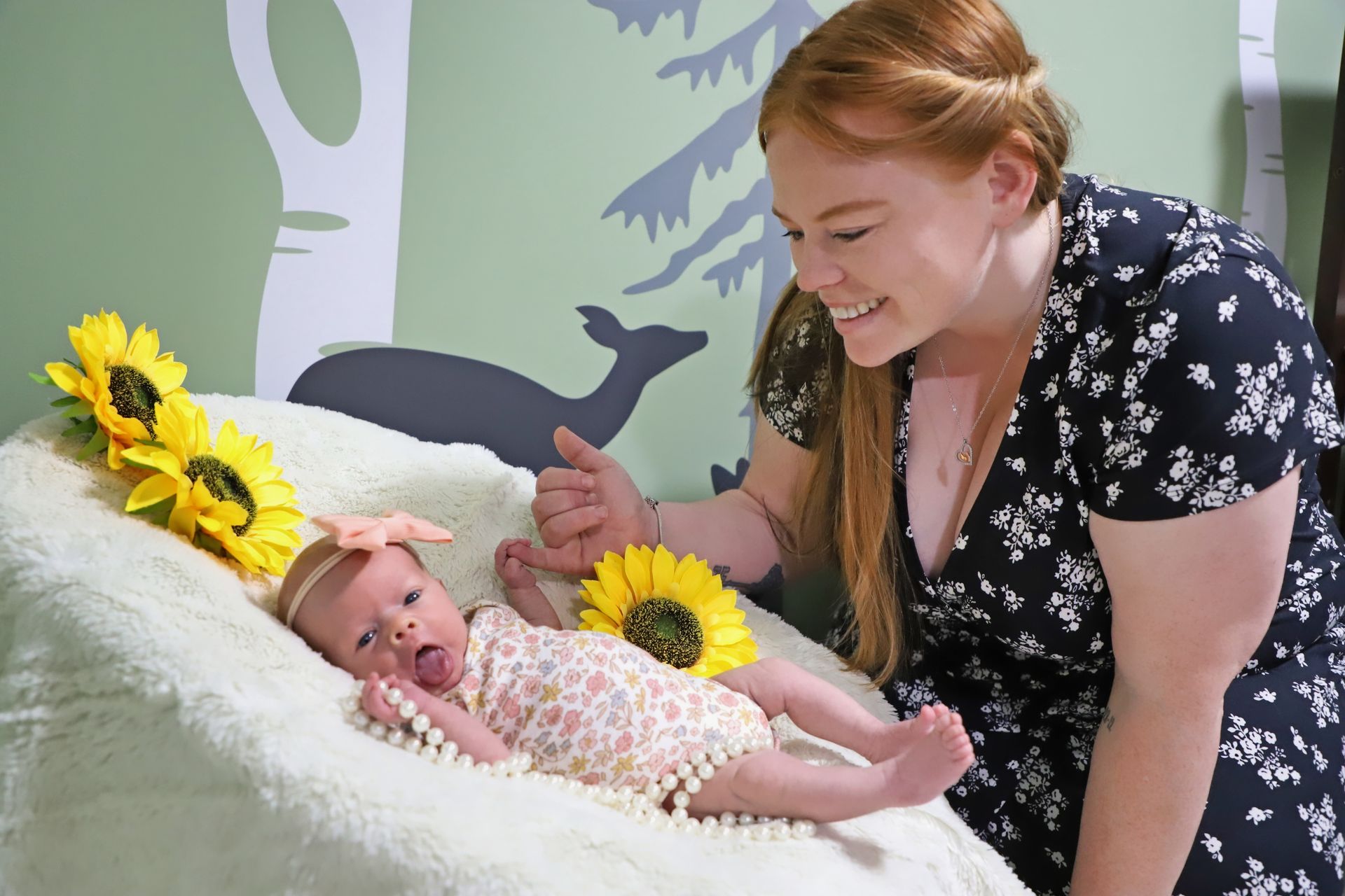 Woman smiles at newborn lying on white cushion with sunflowers. Green and gray wall mural in background.