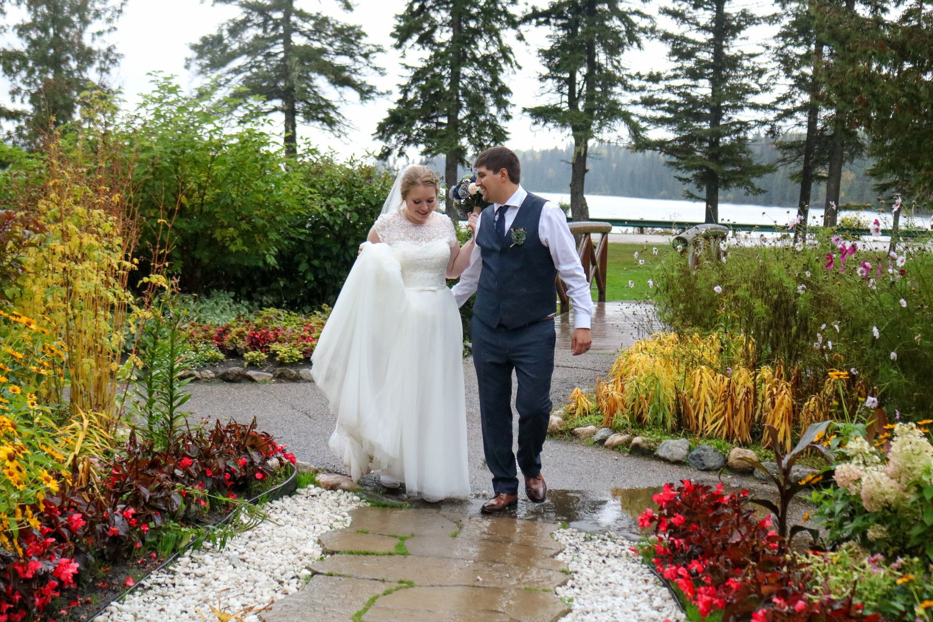 Bride and groom walking hand-in-hand on a path in a garden. She wears a white gown, he wears a vest. Lake in background.