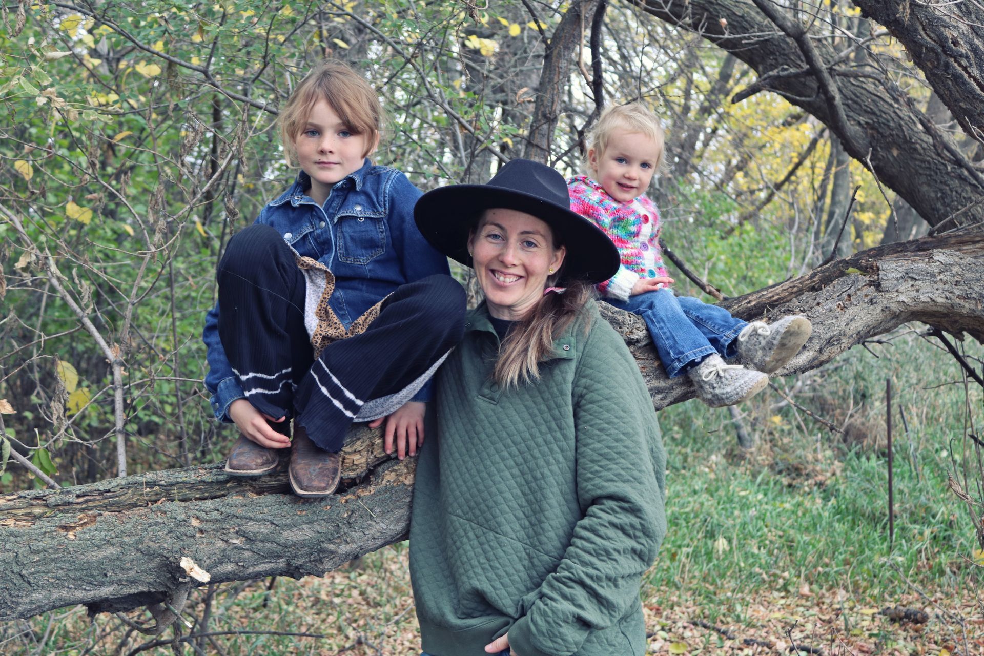 Woman and two children on a tree branch outdoors; woman smiles wearing a hat, kids smile.