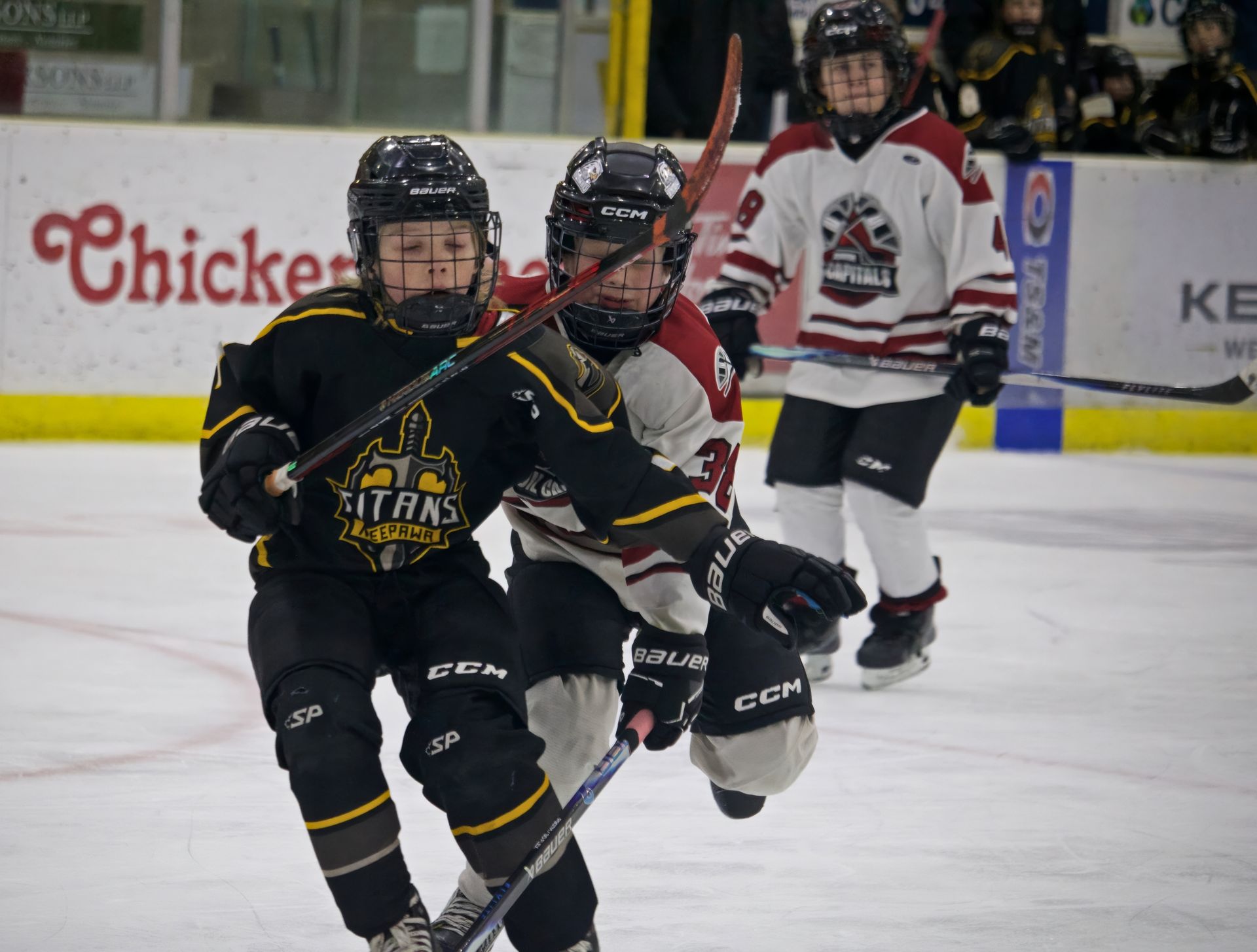 Two youth hockey players in black and white uniforms compete for the puck during a game on an indoor ice rink.