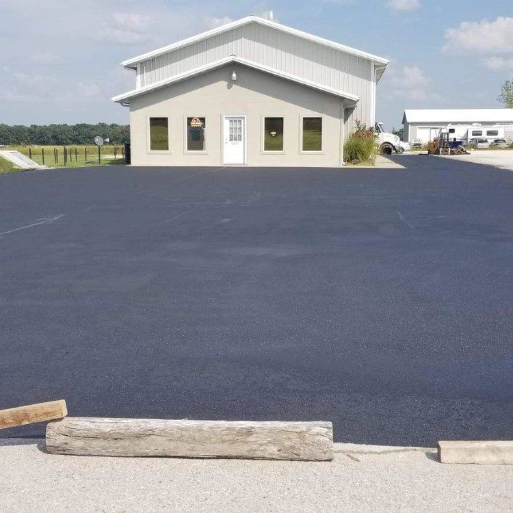 Asphalt parking lot in front of a two-story tan building with a metal roof, clear sunny day.