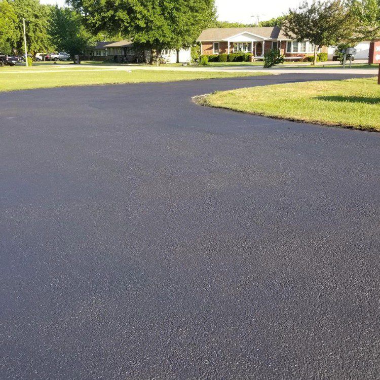 Black asphalt driveway curves in front of a suburban house, bordered by green grass and trees.