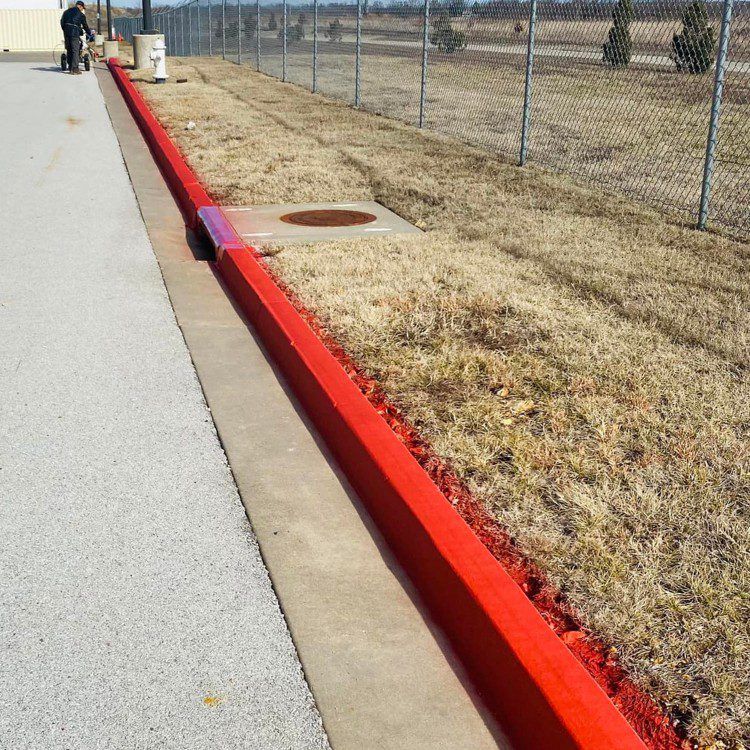 Red curb next to a sidewalk and dry grass, by a chain link fence, person walks by.