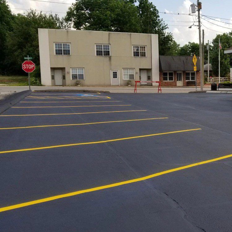 Newly paved parking lot with yellow lines in front of a two-story building. A stop sign is visible on the left.