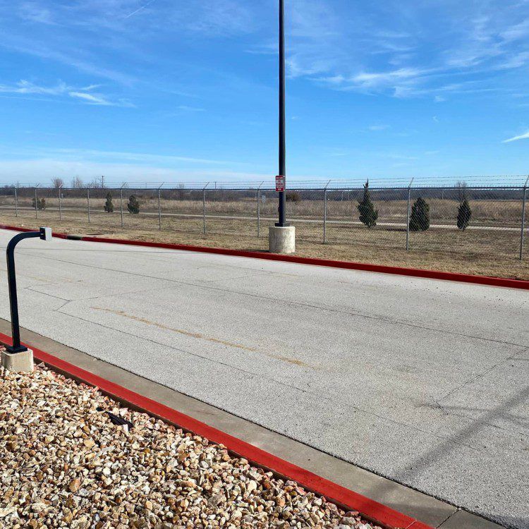 A paved road with red curb, gravel, and a tall black pole, with a wire fence under a blue sky.