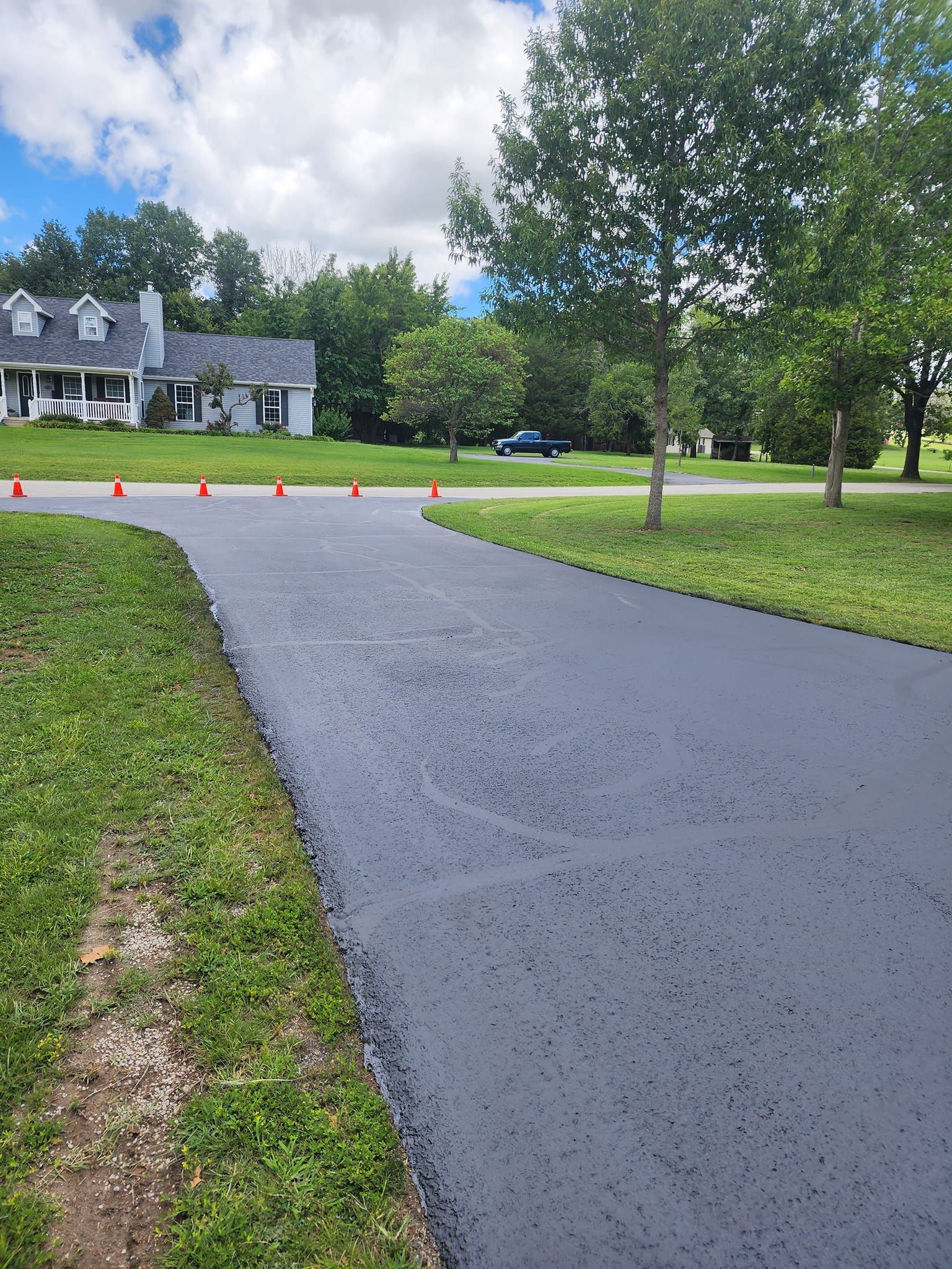 Newly paved asphalt driveway with orange traffic cones, leading to a grey house with green lawn and trees.