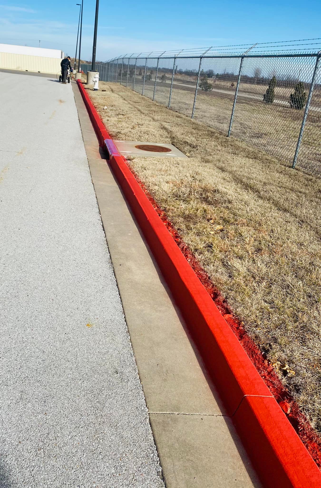 Red curb, gravel walkway, chain-link fence, bright blue sky.