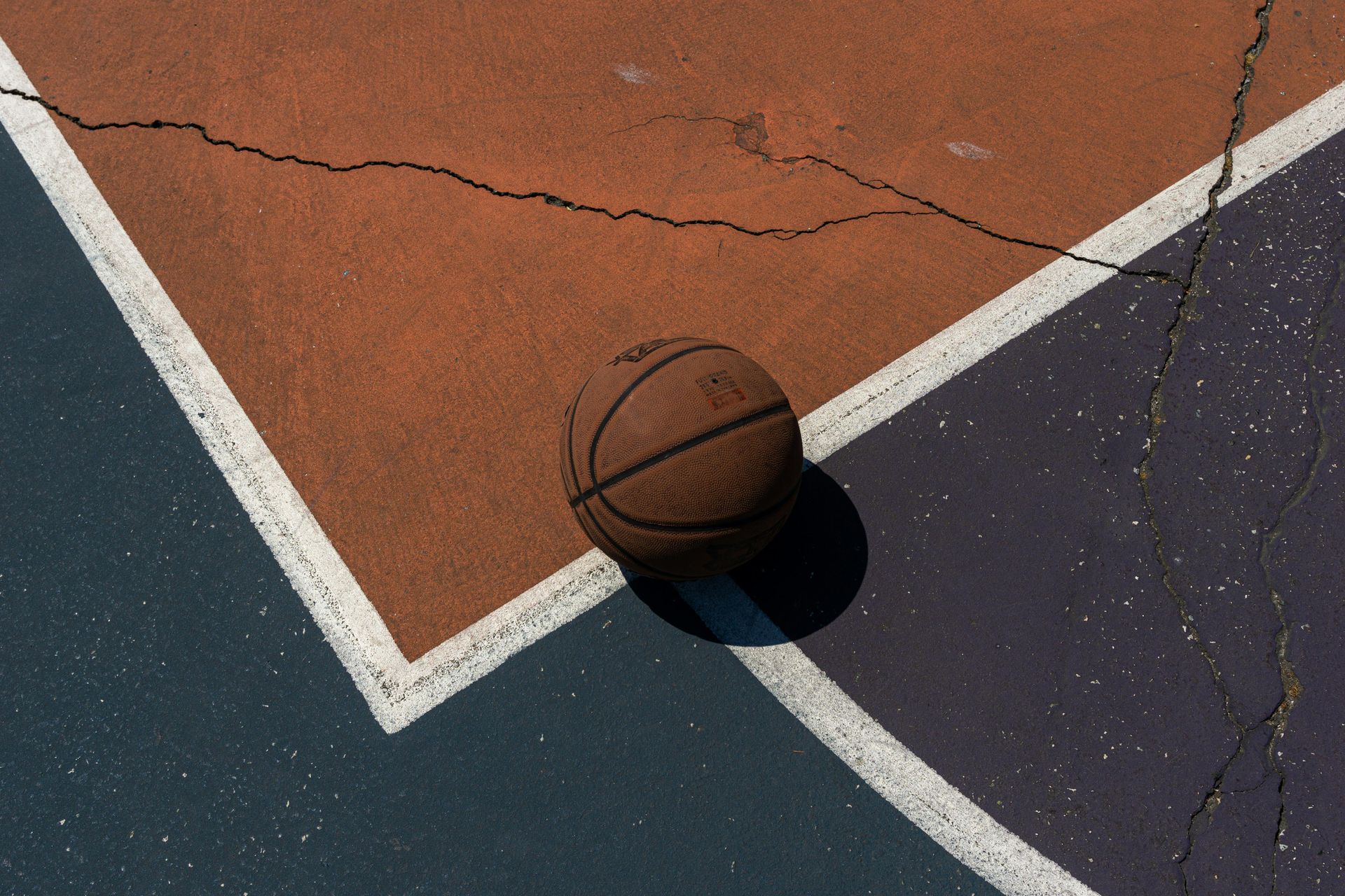 Basketball on cracked court, corner of the court marked with white lines, brown and blue surfaces.