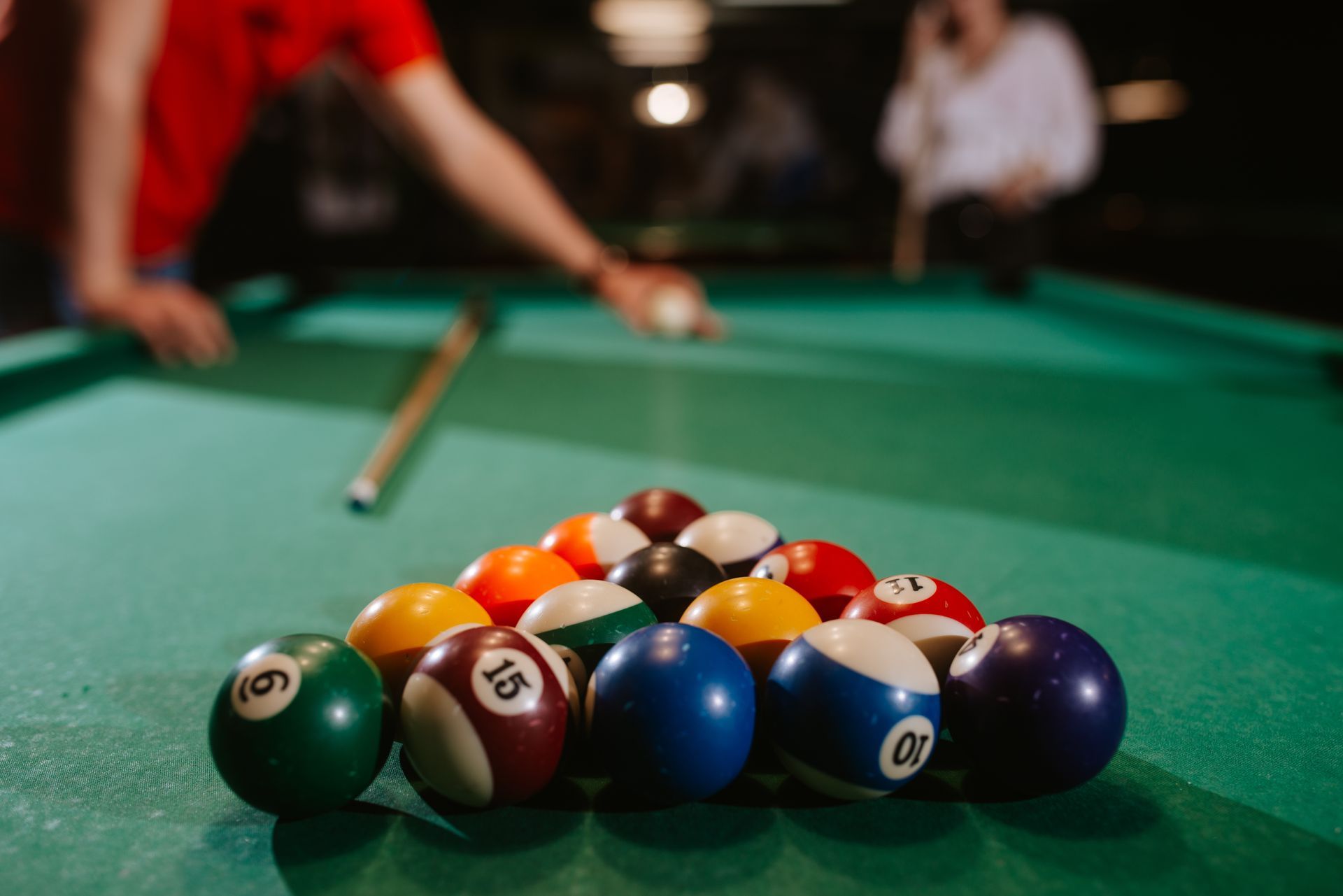 Pool table with balls scattered on green felt, two people playing in a dimly lit setting.