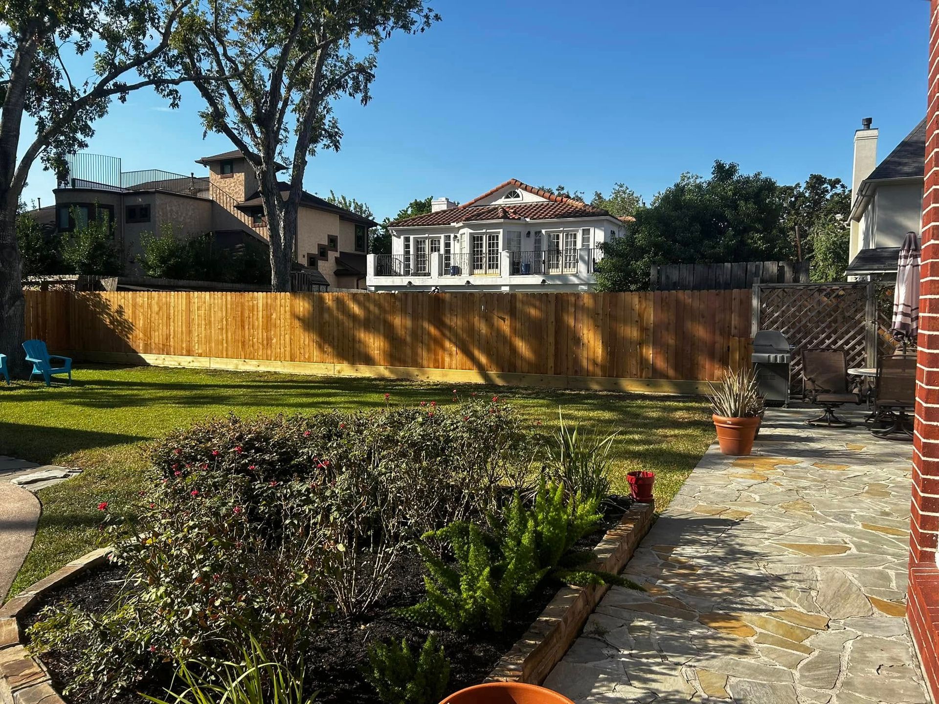 Backyard with a brown wooden fence, green lawn, and two-story house in the background under a blue sky.