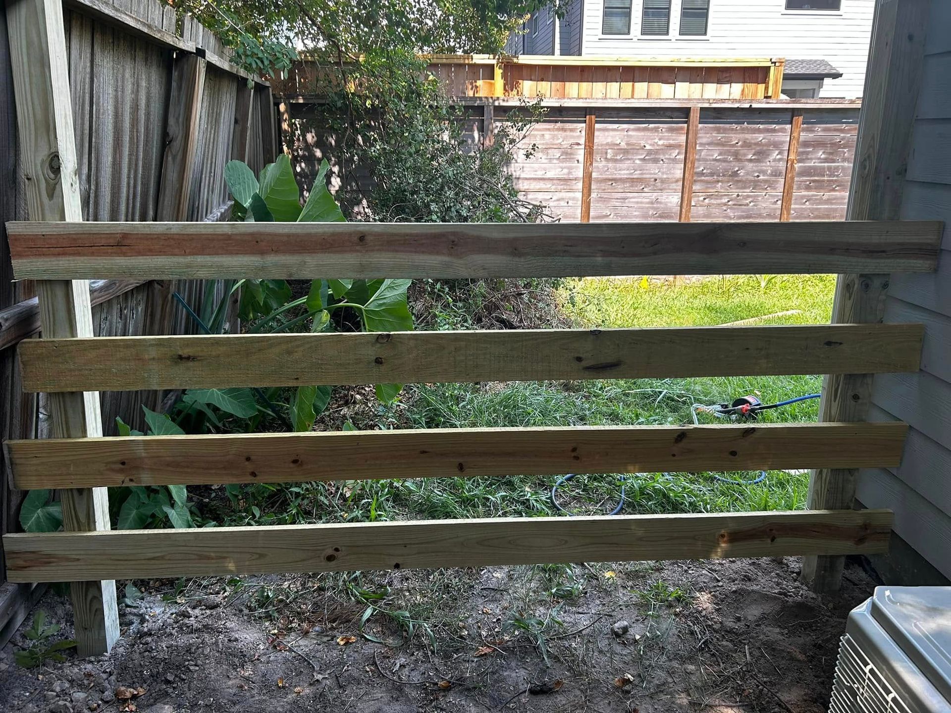 Wooden fence being constructed, with three horizontal planks between supporting posts. Green foliage visible behind.