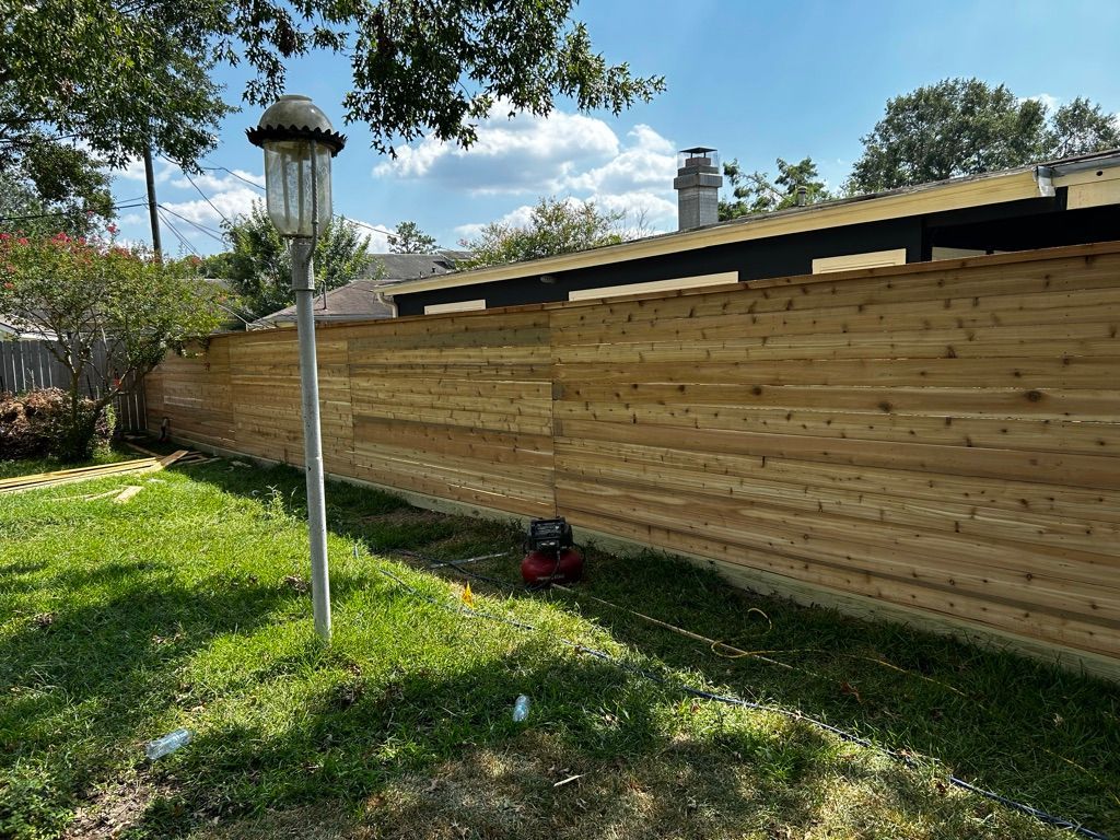 A wooden privacy fence in a grassy yard, a lamp post, a red lawn mower, and trees under a blue sky.