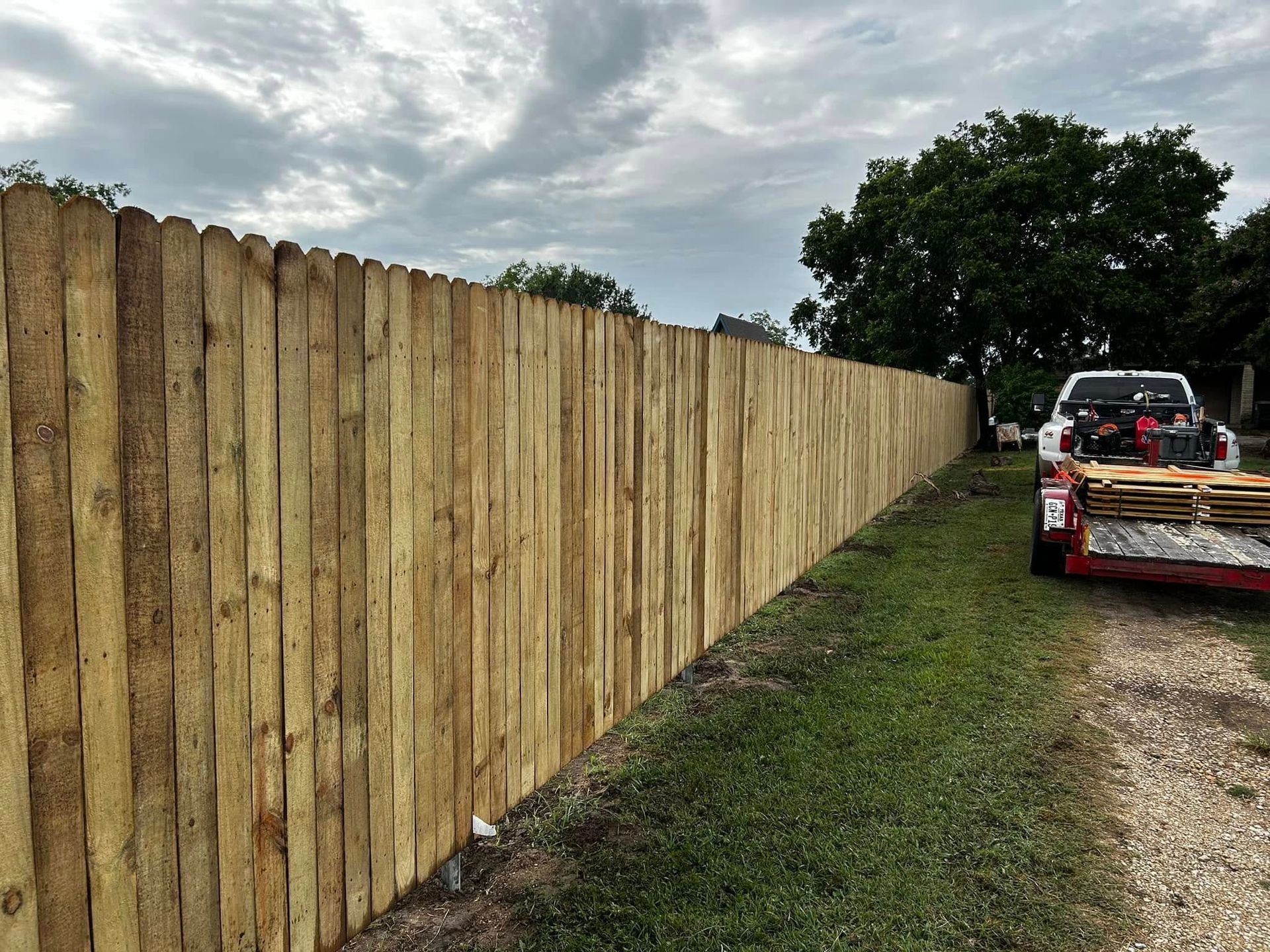Wooden privacy fence next to a grassy area, with a truck parked nearby under a cloudy sky.