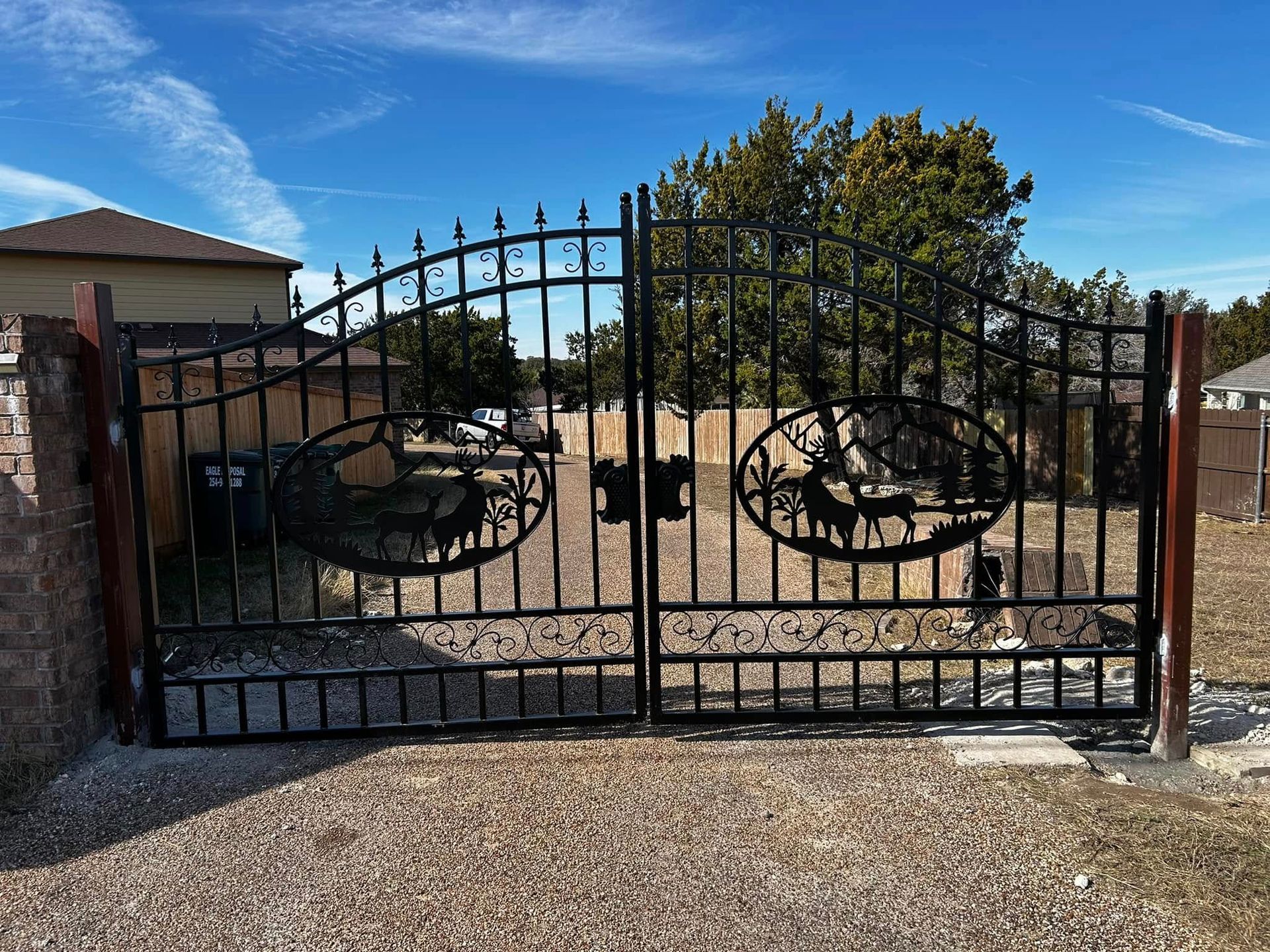 Black wrought iron gate with deer silhouettes, set against a blue sky, gravel driveway.