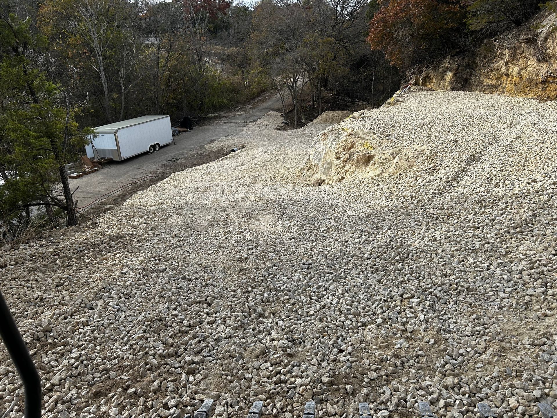 Gravel-covered slope with a white trailer at the top. Trees and a rocky embankment flank the graded area.