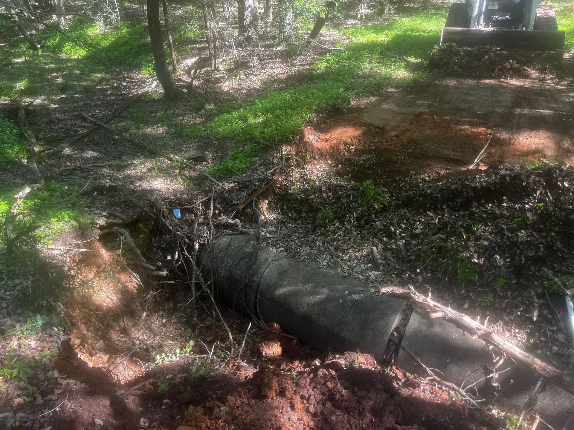 Black pipe partially buried in dirt, next to a grave in a wooded area.
