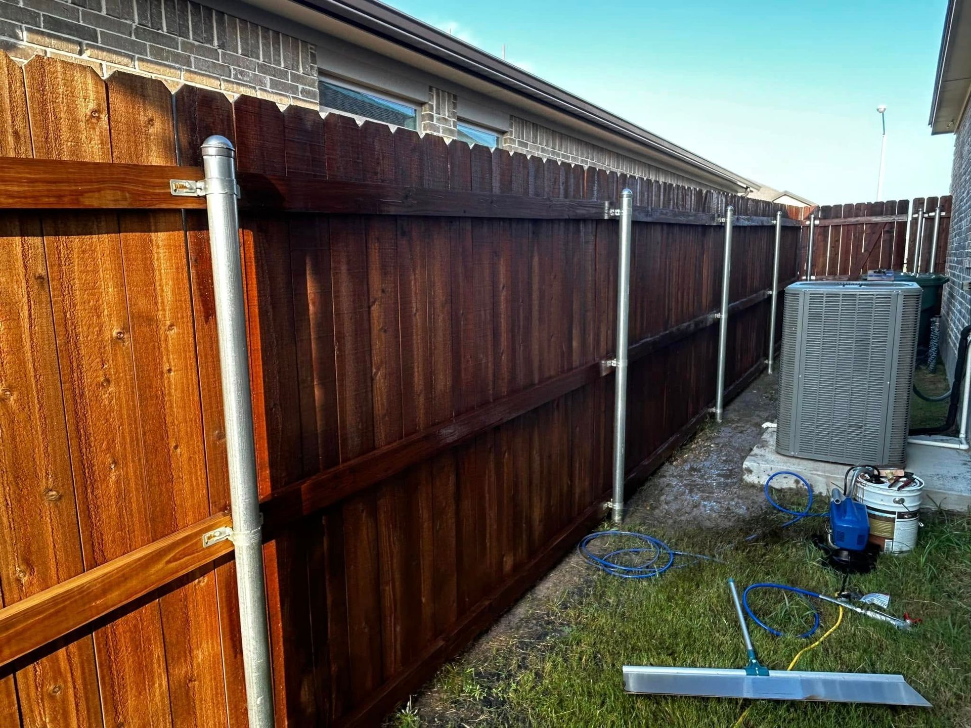 Wooden fence stained dark brown, with metal poles for support, next to a building and air conditioning unit.