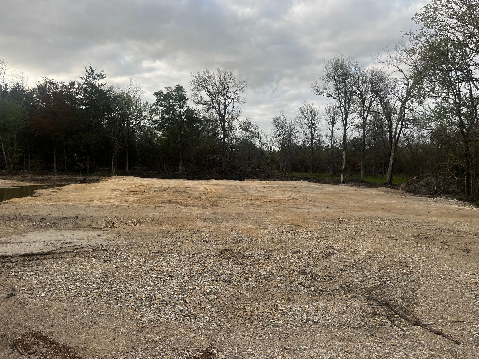 Open, gravel-covered clearing surrounded by bare trees under a cloudy sky.