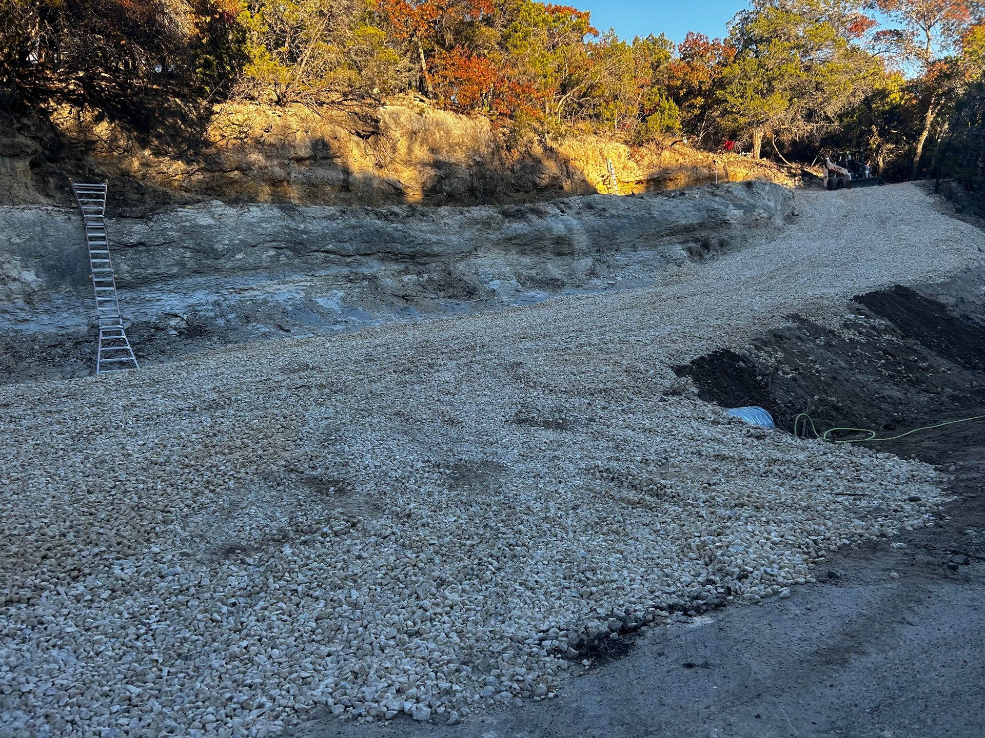 Gravel-covered area with a cut bank on a hillside. A ladder stands on the bank. Trees in the background.
