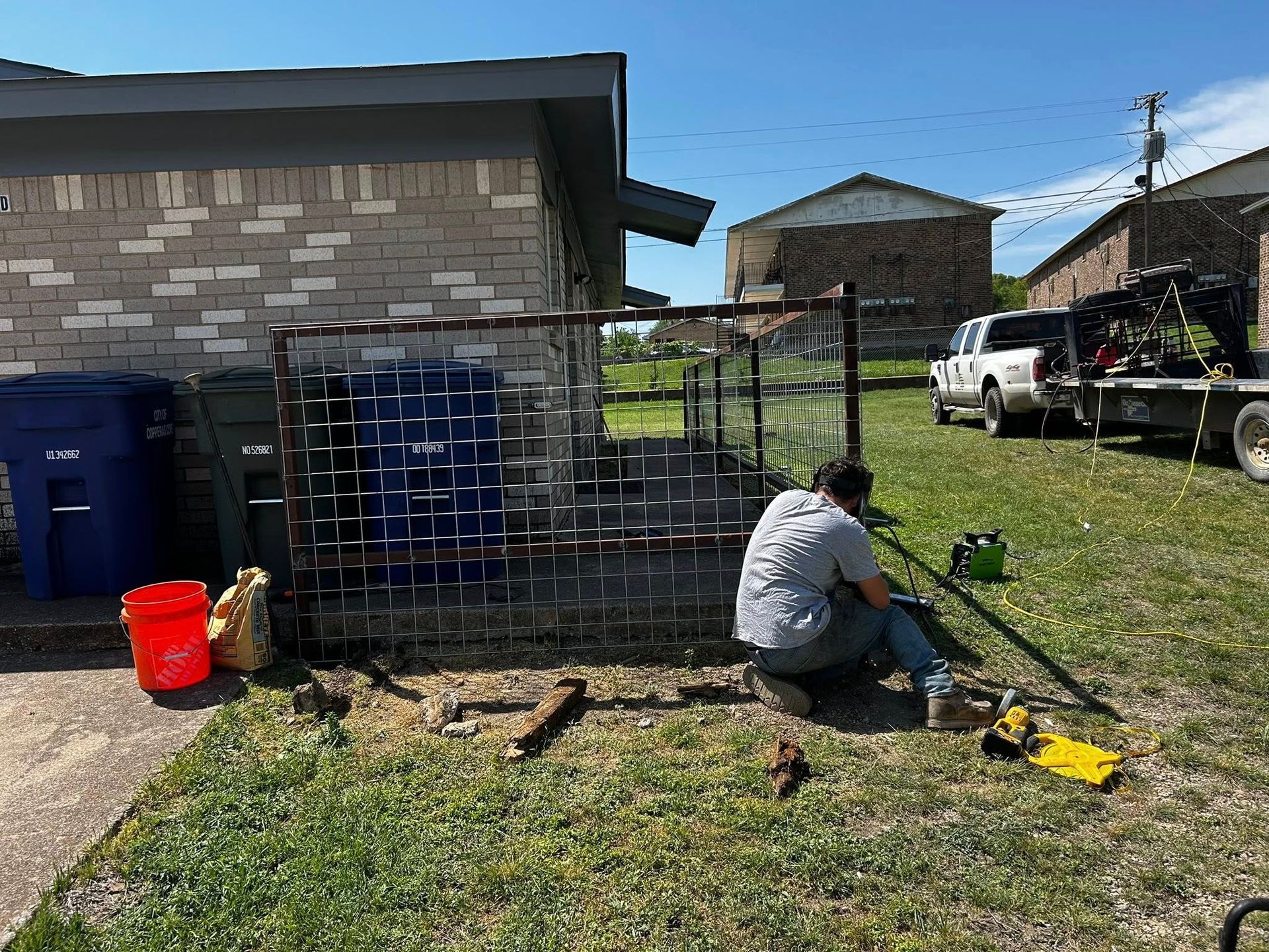 Person repairing a fence near a building. A truck and trash cans are visible in the background on a sunny day.