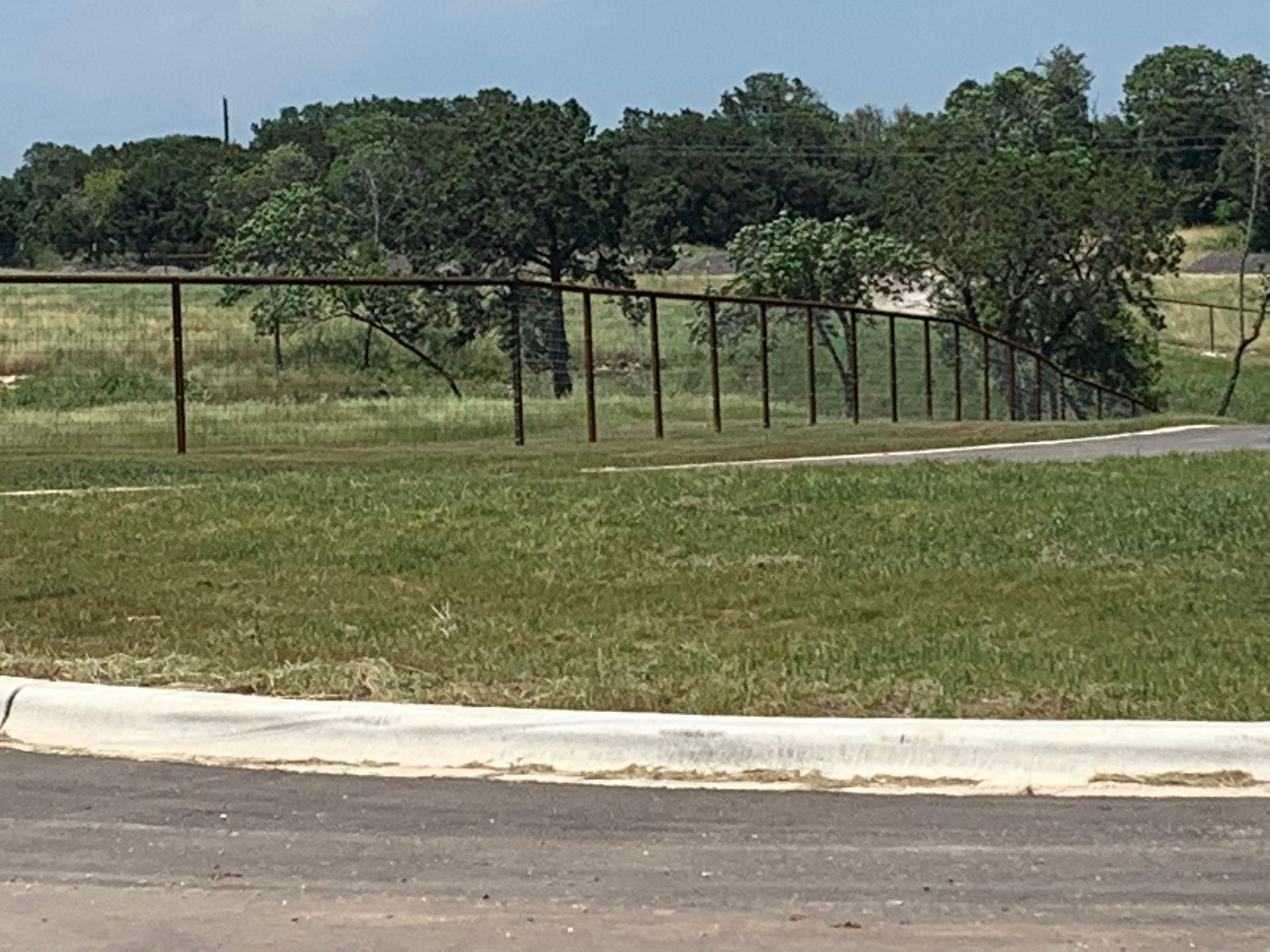 Chain link fence bordering green grass and trees, with a road in the foreground.