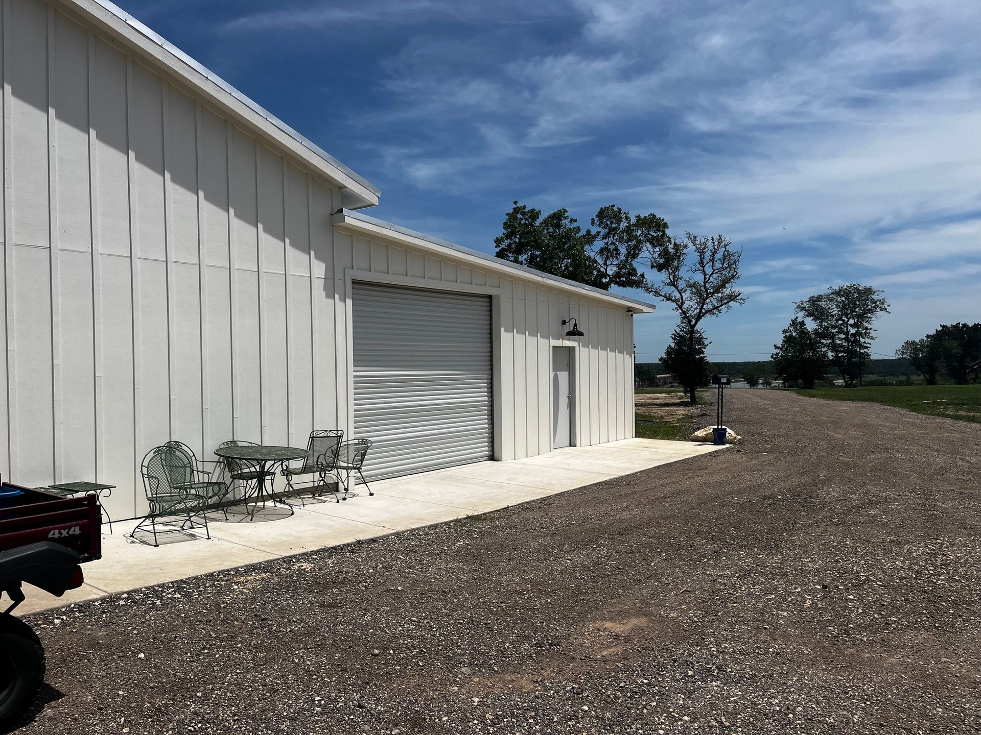 White building with a roll-up door, side entrance, and small patio set on a gravel driveway under a blue sky.