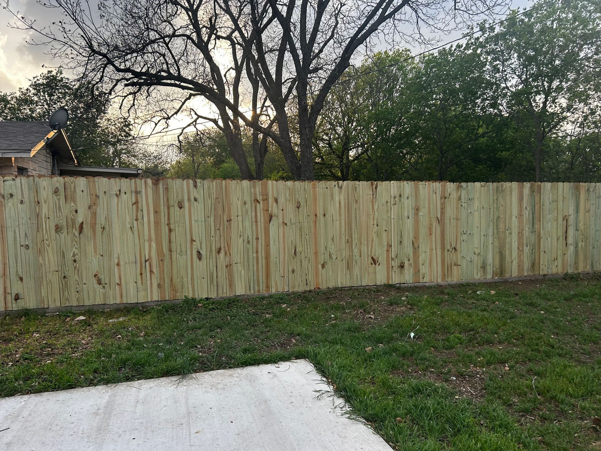 Wooden fence in backyard, with concrete slab in foreground, trees and cloudy sky in background.