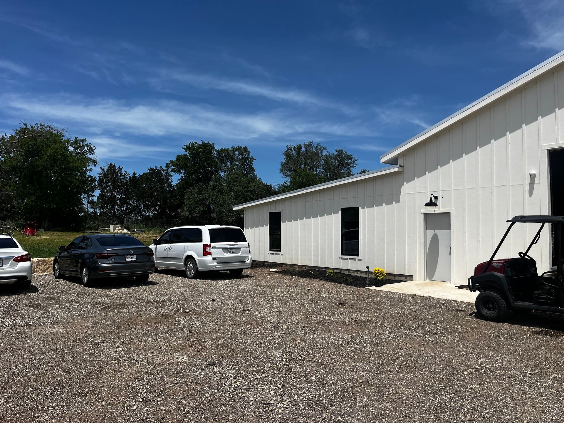 White building with parked cars on a gravel lot under a blue sky with some trees.