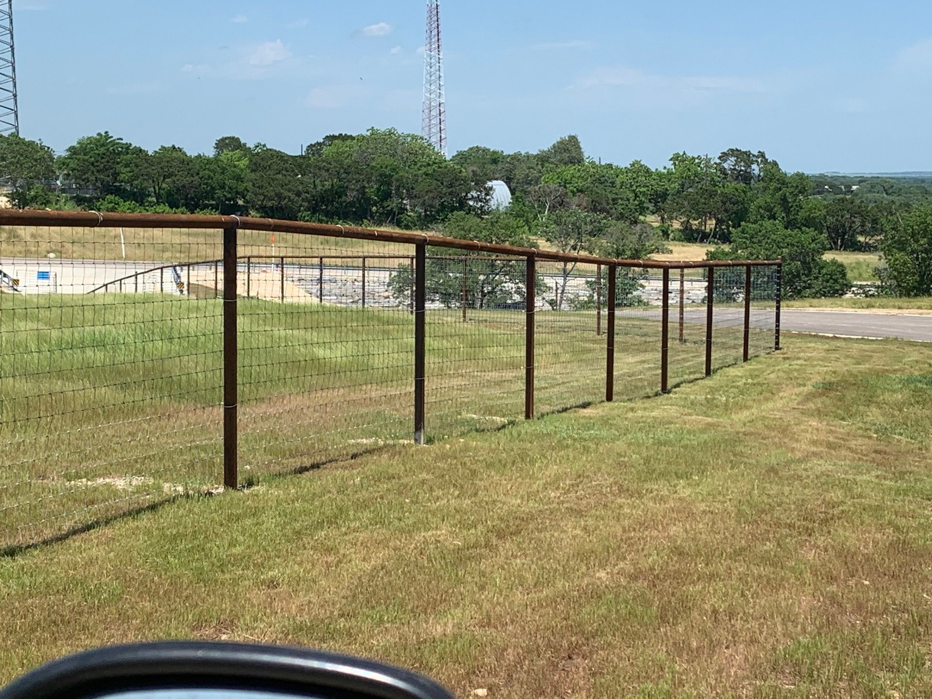 Brown metal fence on a grassy hill; trees and a blue sky in the background.
