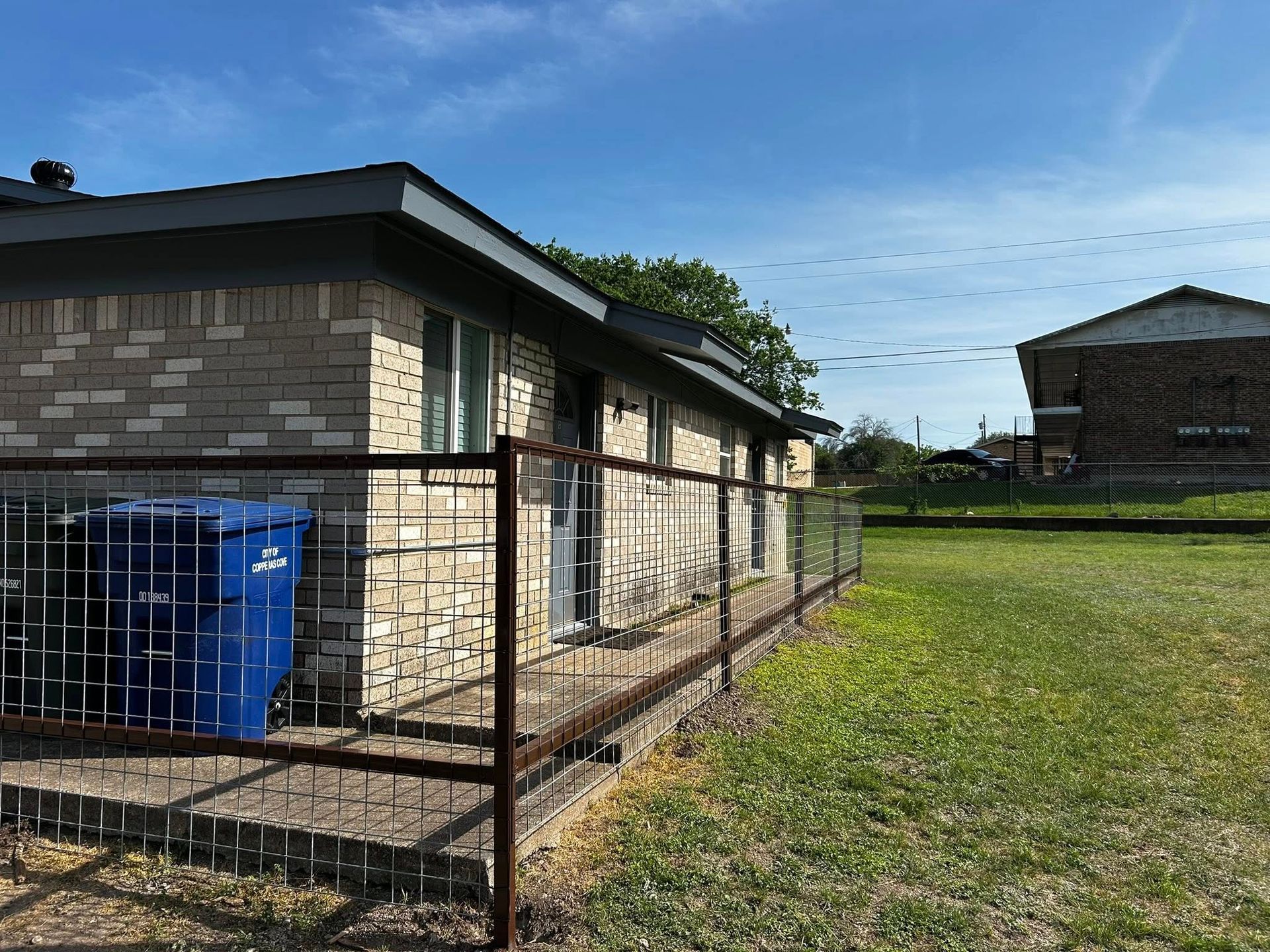A brick house with a metal fence and blue trash bin on a grassy lawn under a blue sky.