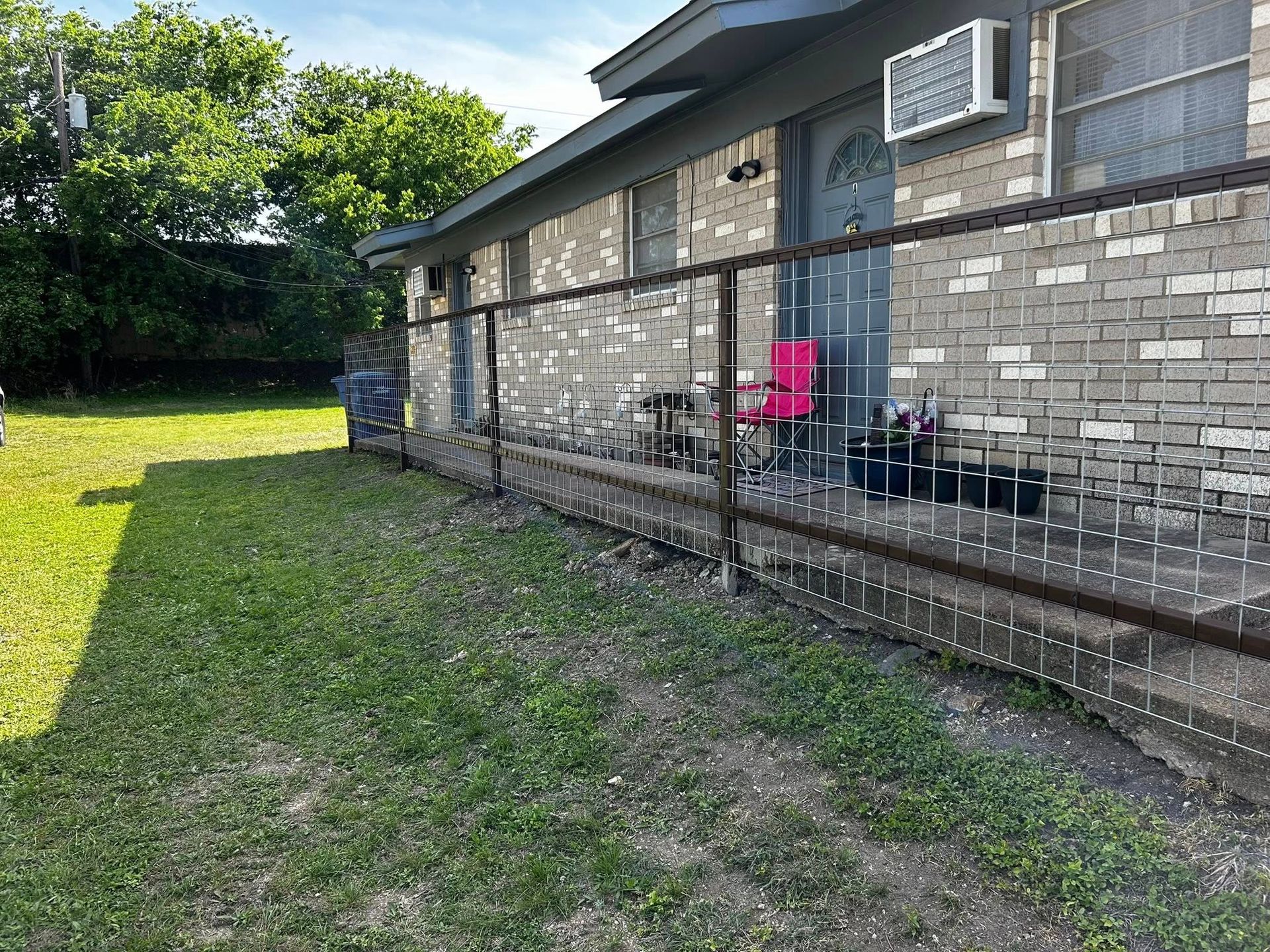 Brick building with a metal fence, green lawn, a pink chair, and an air conditioner.
