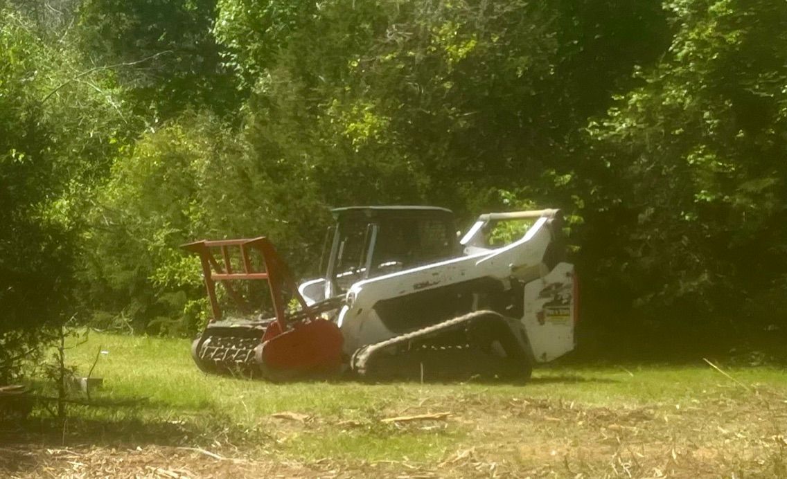 White tracked skid-steer with a mulching attachment in a grassy area, surrounded by trees.
