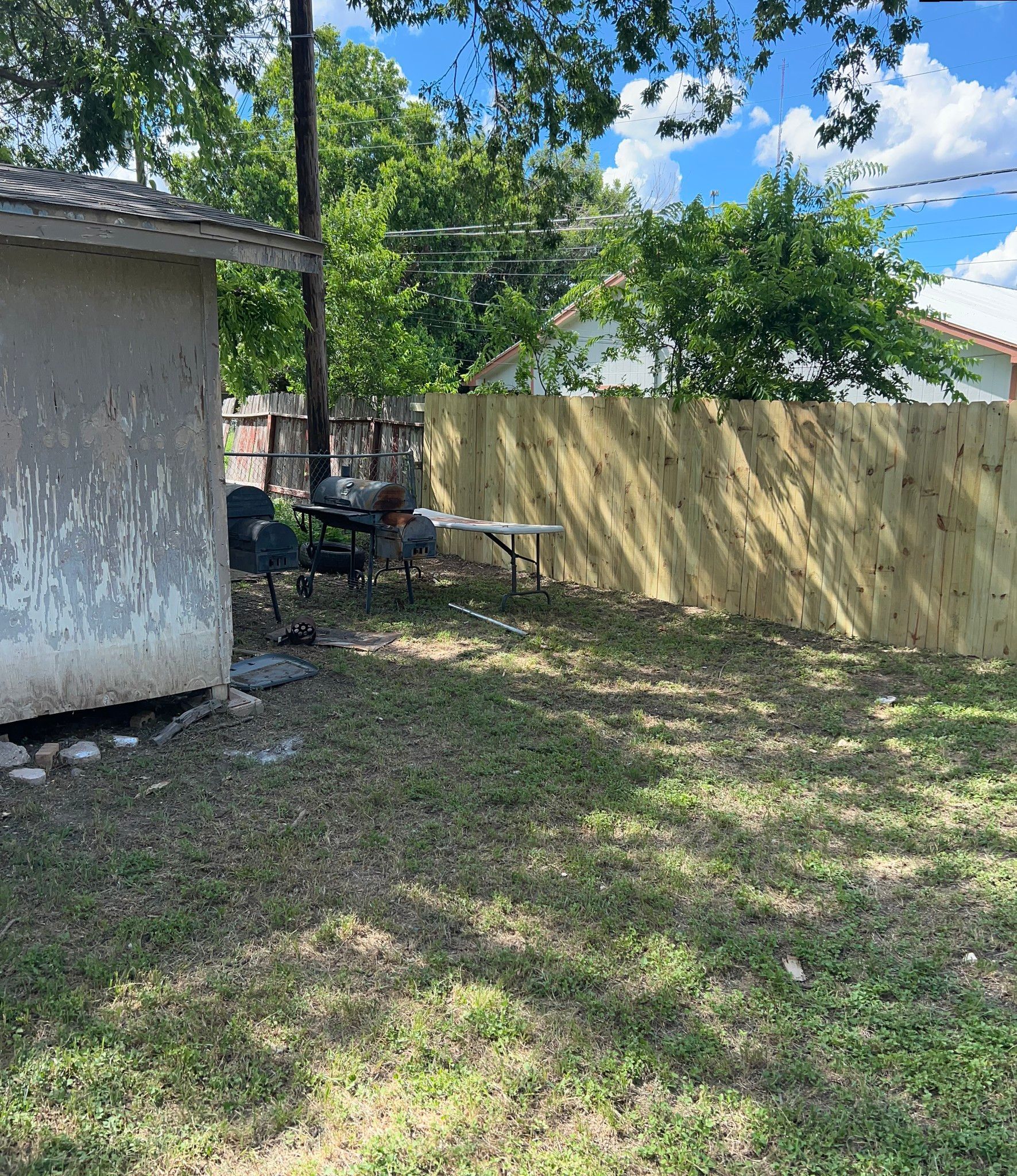 Backyard with new wooden fence, shed, table, and dead grass under a blue sky.
