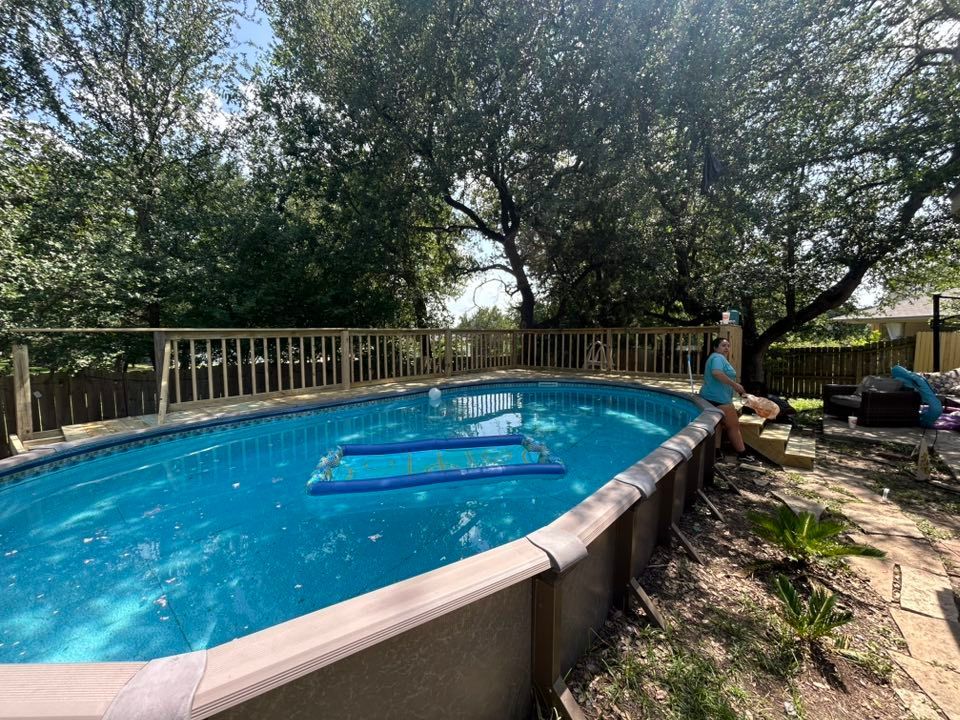 Oval above-ground pool with blue water, surrounded by a wooden deck and trees. A person sits nearby.