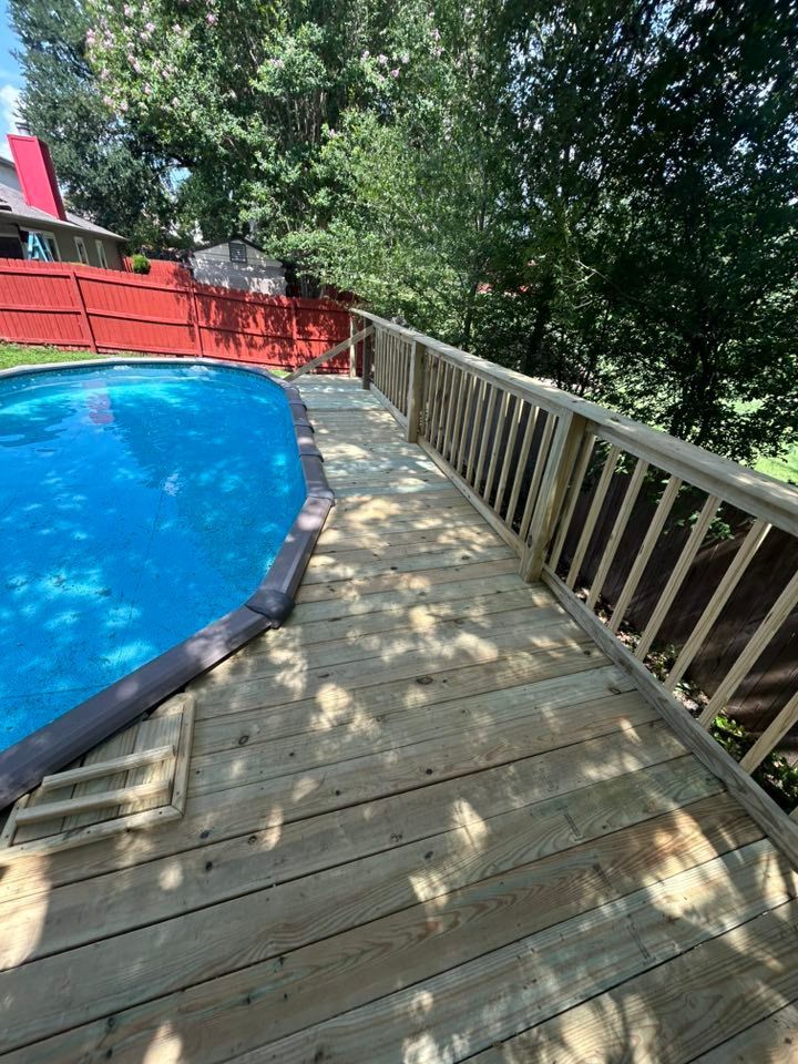 Wooden deck surrounding a blue pool, with railing, near trees and red fence.