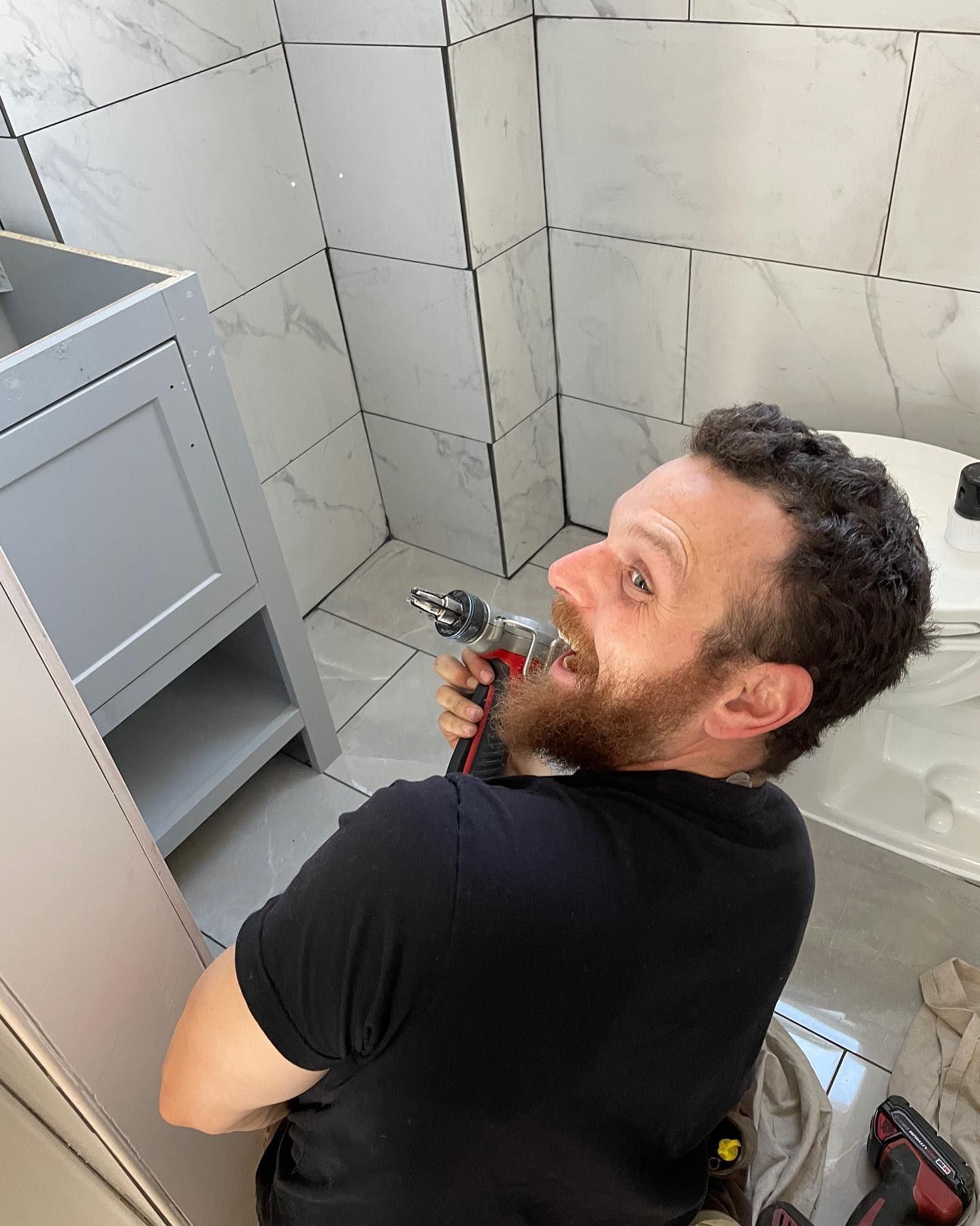 A man with a beard is using a drill in a bathroom.