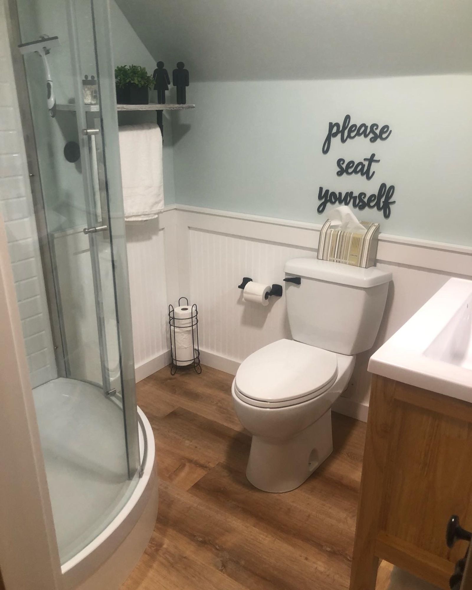 A bathroom with a toilet , sink , shower and a sign on the wall that says `` please seat yourself ''.