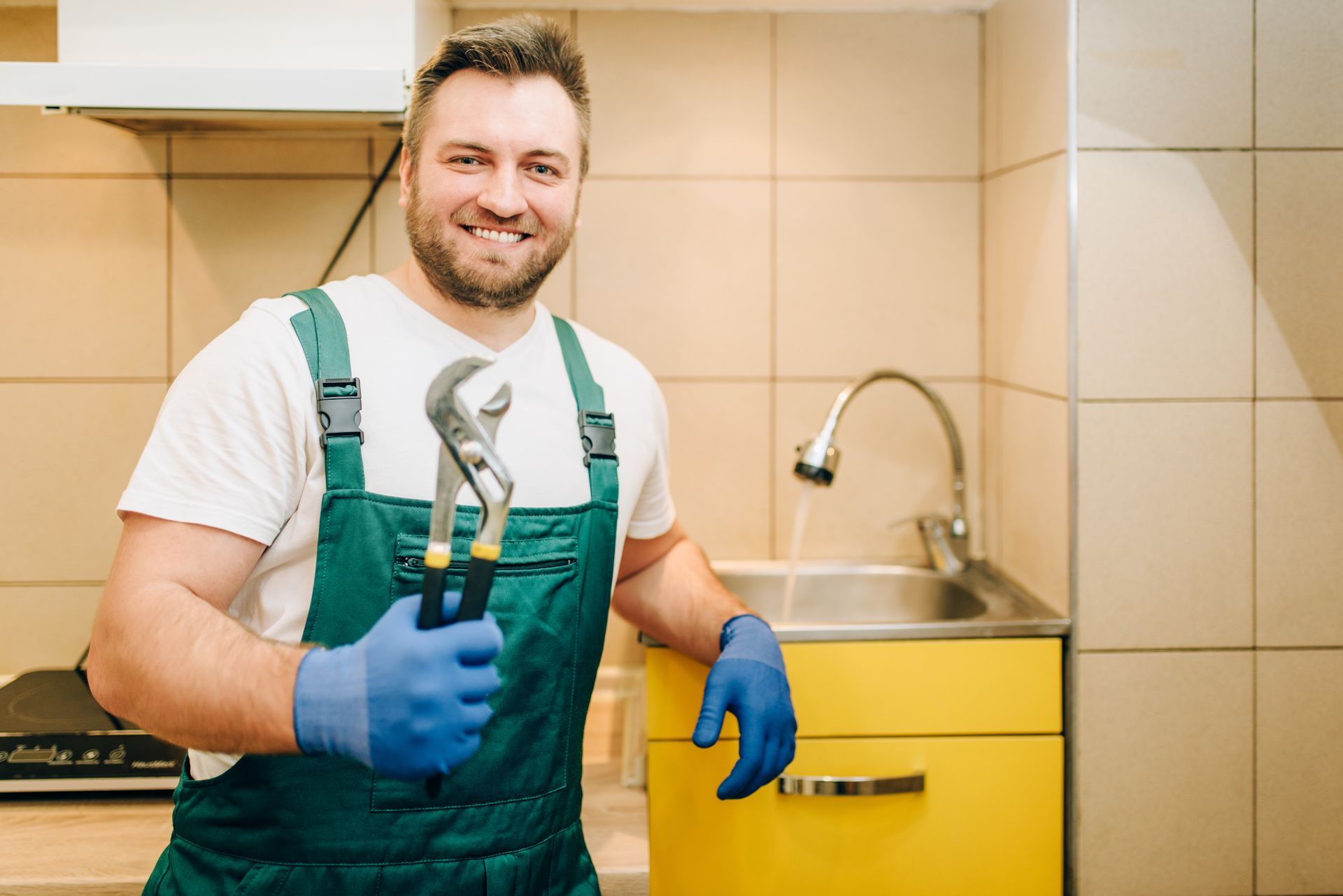 A man in overalls is holding a pair of wrenches in a kitchen.