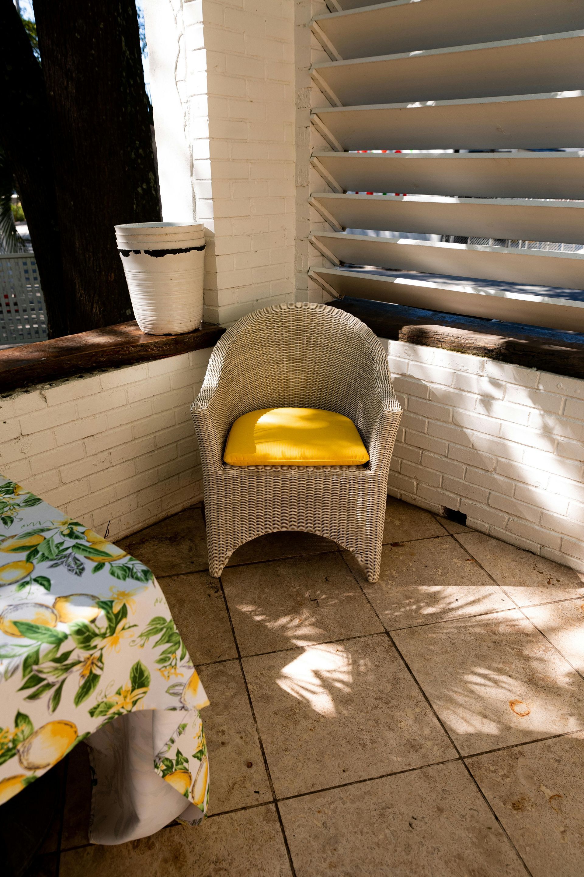 Wicker chair with yellow cushion on a tiled patio, next to a table with lemon-print tablecloth and a slatted wall.