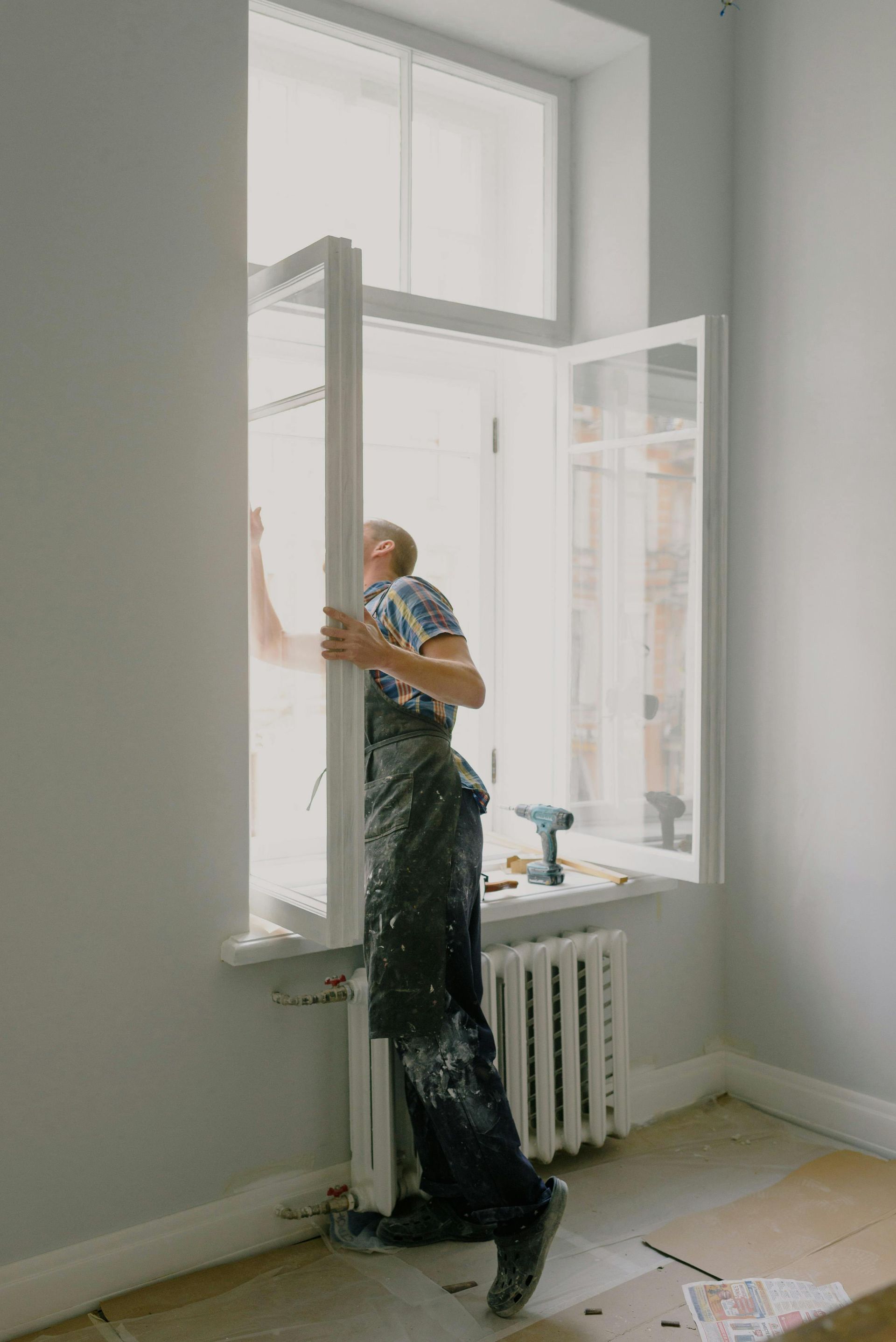 Man in overalls installing a window frame in a room with gray walls.
