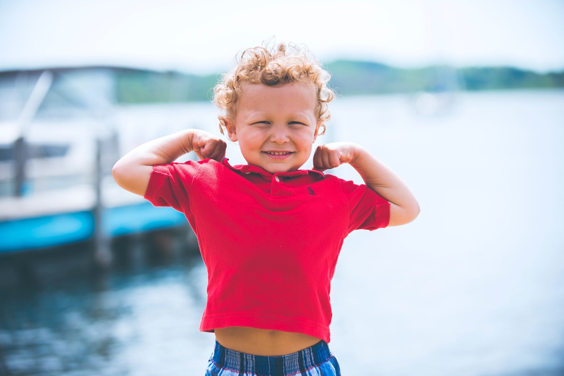 A young boy in a red shirt is flexing his muscles in front of a body of water.