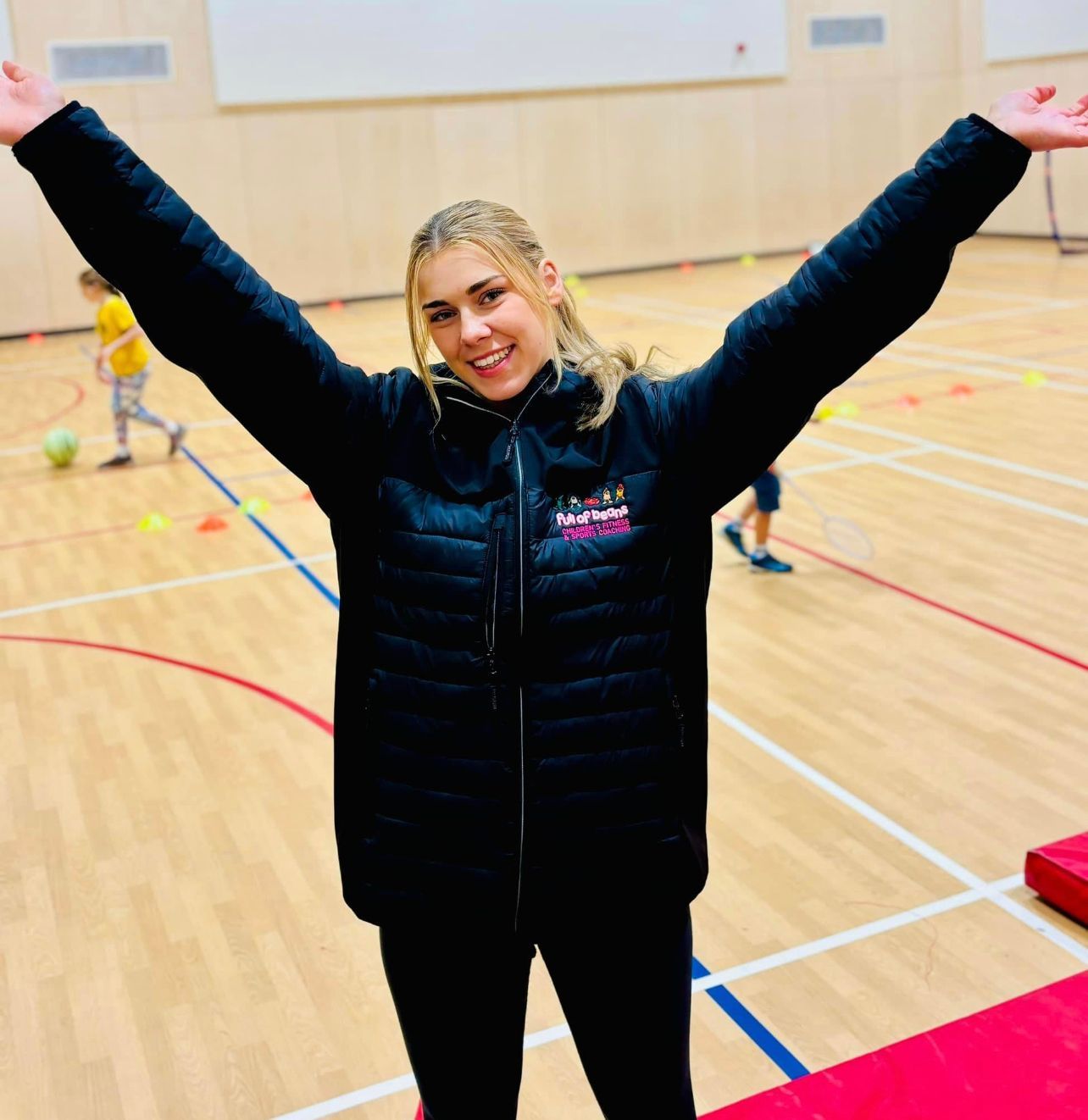 A woman in a black jacket is standing in a gym with her arms outstretched.