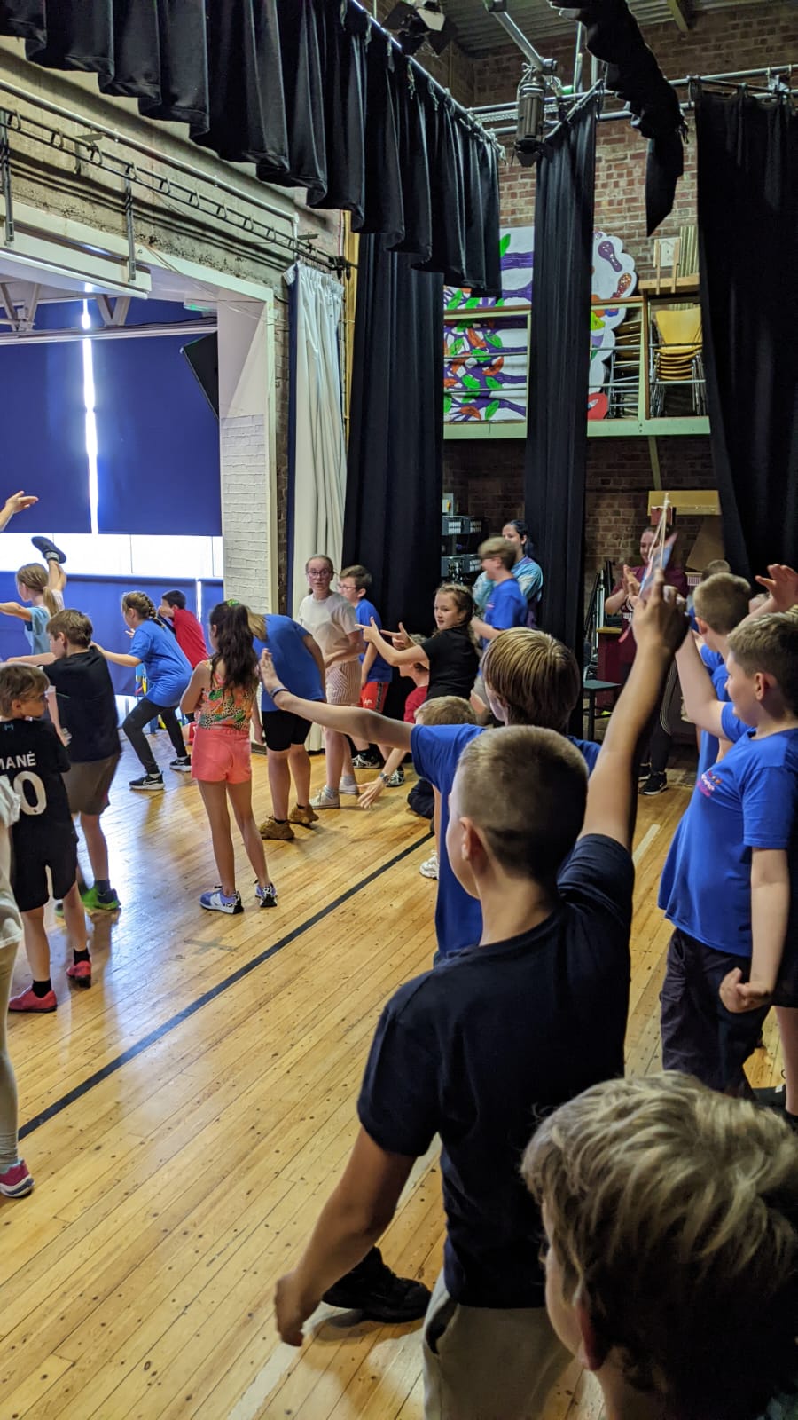 A group of children are standing in a gym raising their hands.