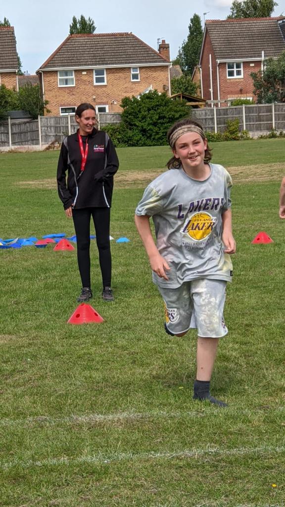 A young boy is running on a field with a woman standing behind him.